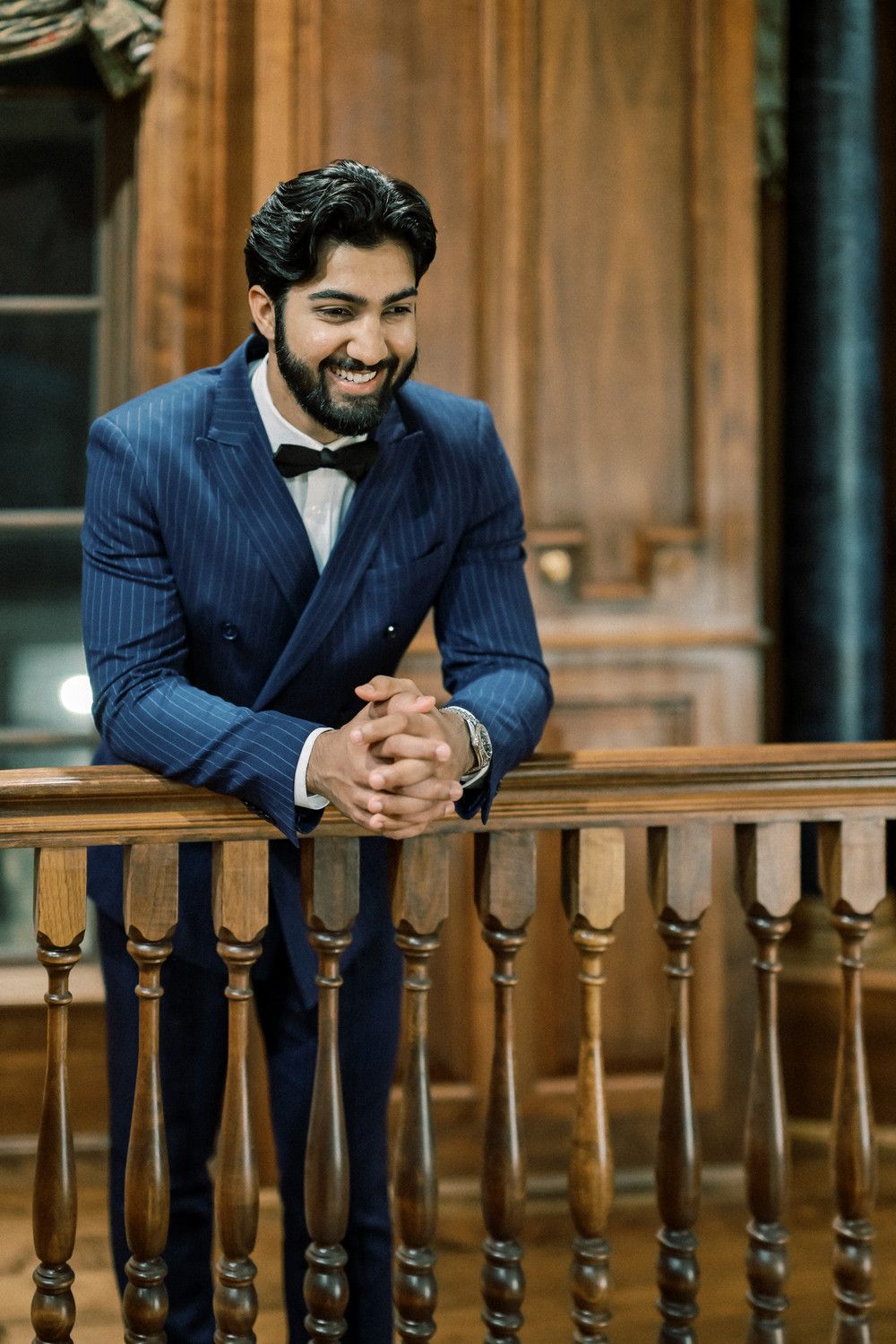 Smiling man in navy pinstriped suit leans on wooden balcony railing.