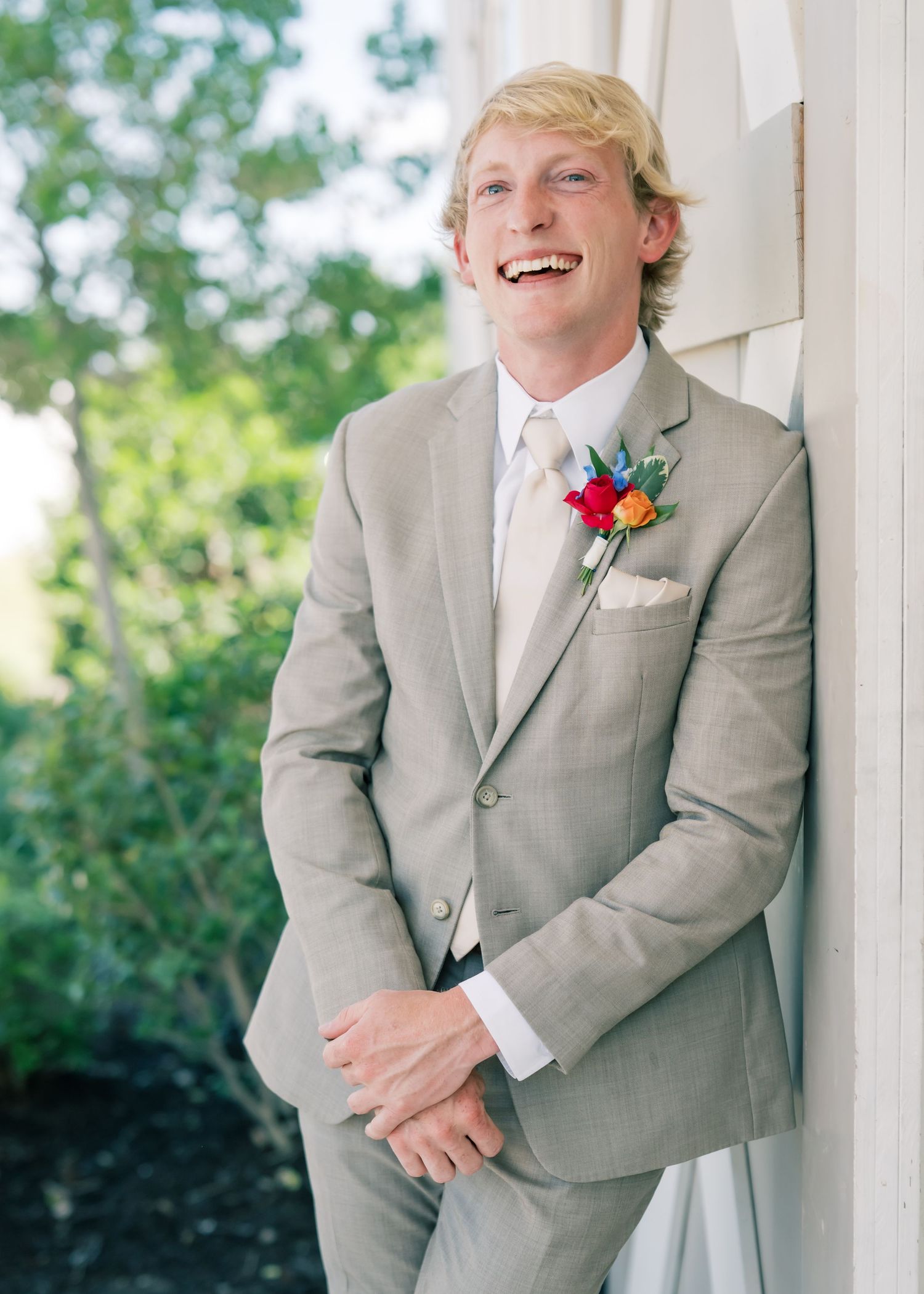 Smiling groom in light gray suit with colorful boutonniere leaning against white wall outdoors.
