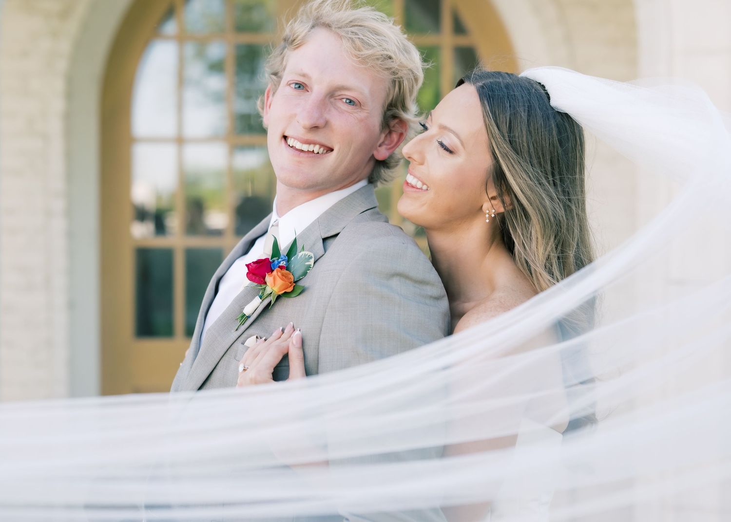 A smiling groom in a gray suit with a colorful boutonniere stands with his bride in a white veil outdoors.