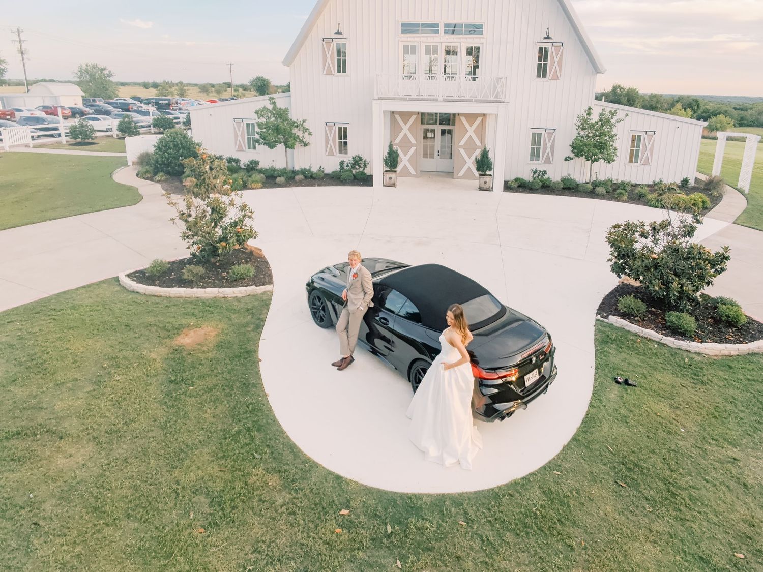 Aerial view of a bride and groom posing beside a black luxury car in front of a white barn wedding venue.