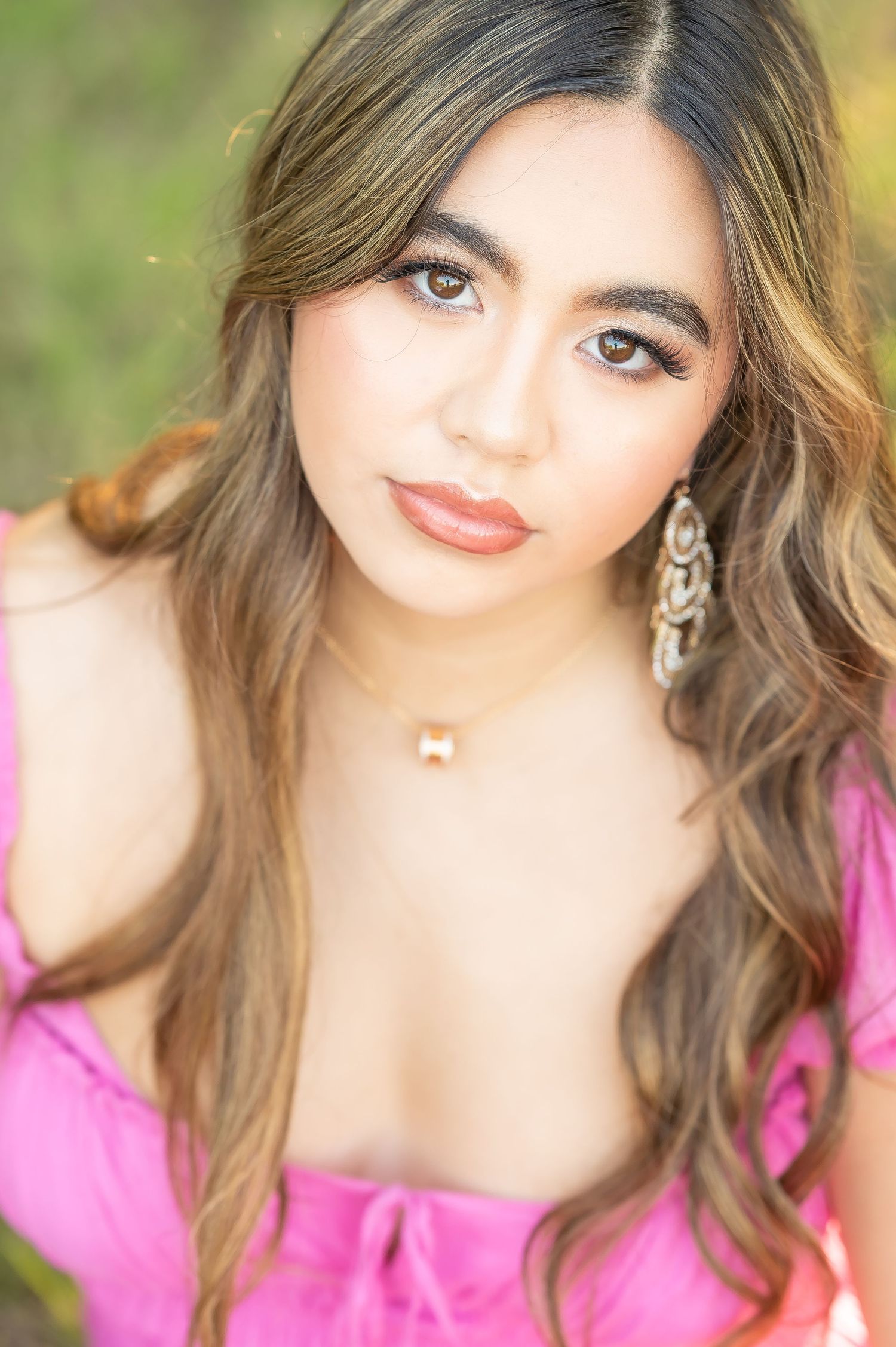 Portrait in pink dress with wavy brown hair, dangling earrings and natural makeup against blurred green background.