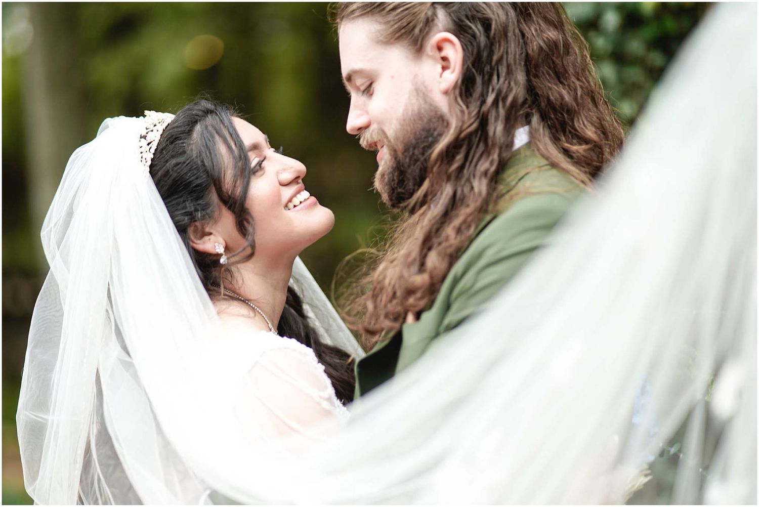 Bride in white veil and groom with long hair share a joyful moment, smiling at each other outdoors.