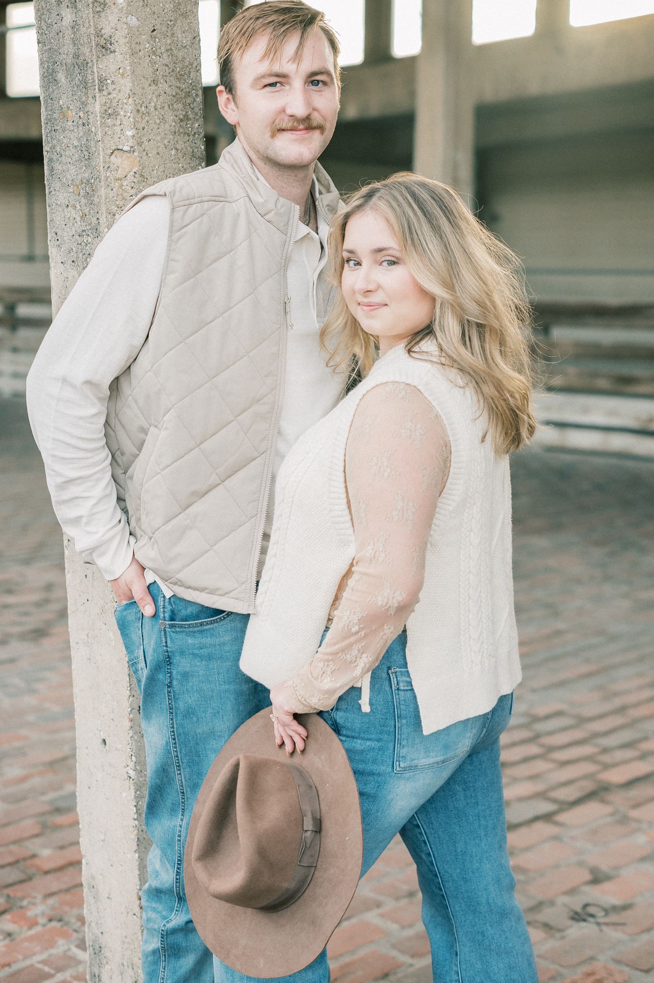 Couple in light colored clothing and western wear posing playfully with a brown hat.