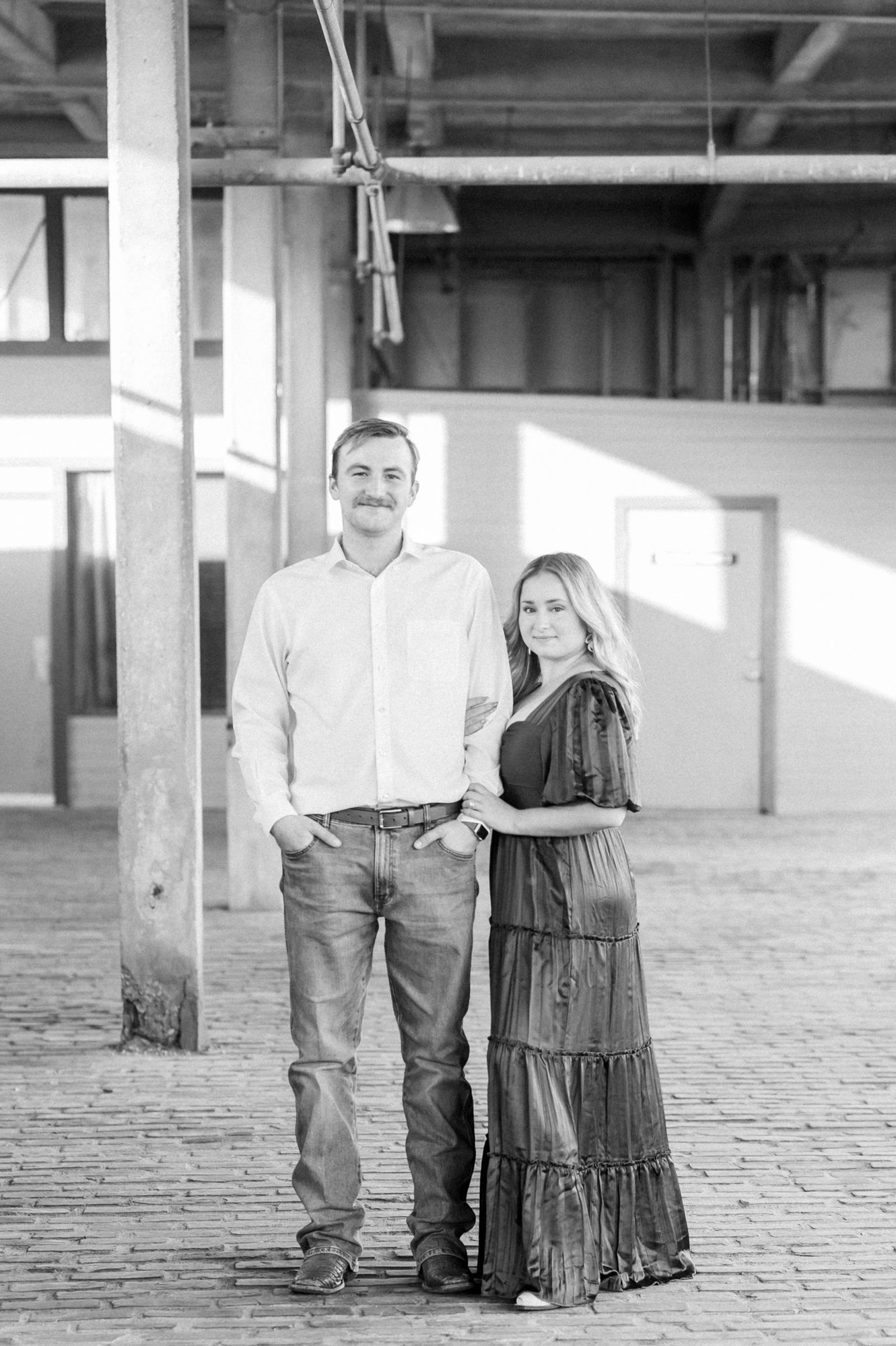 A couple poses together in an industrial space with brick floors and columns, with the woman wearing a long tiered dress.