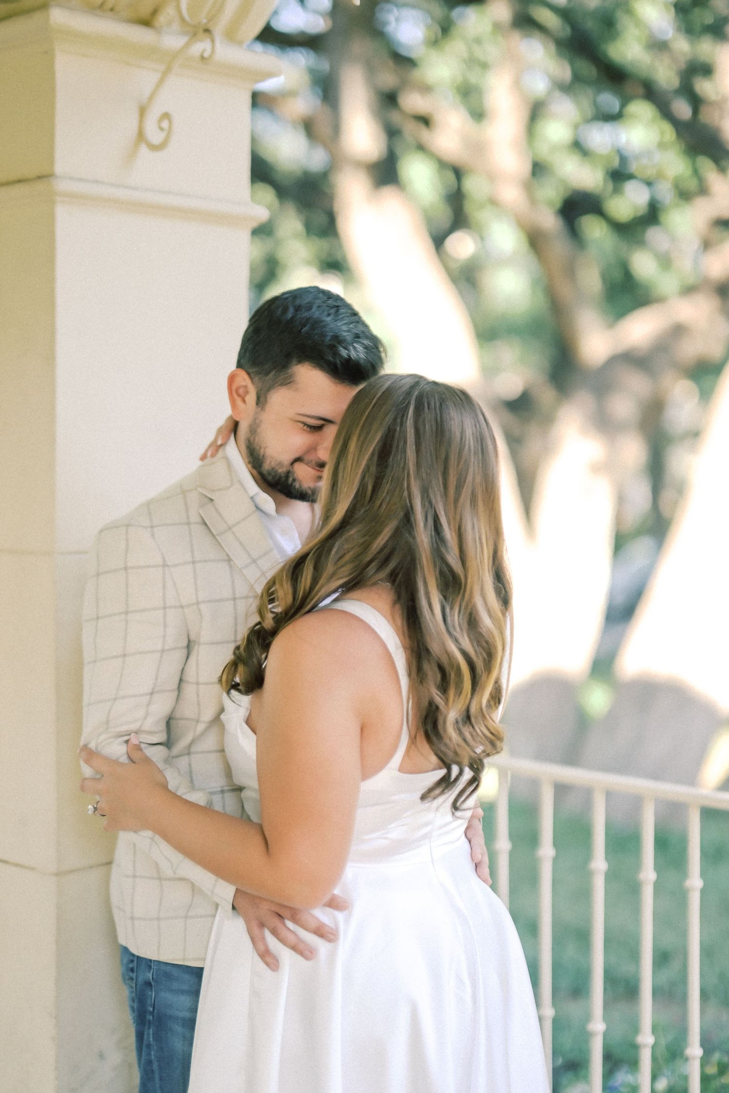 Couple embracing affectionately on a sunlit porch, the woman in a white dress and the man in a plaid blazer, with trees in the background.