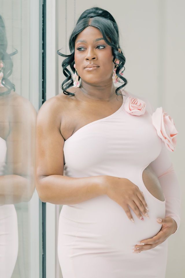 Pregnant woman in pink one-shoulder dress with rose detail, cradling baby bump, standing by mirror.