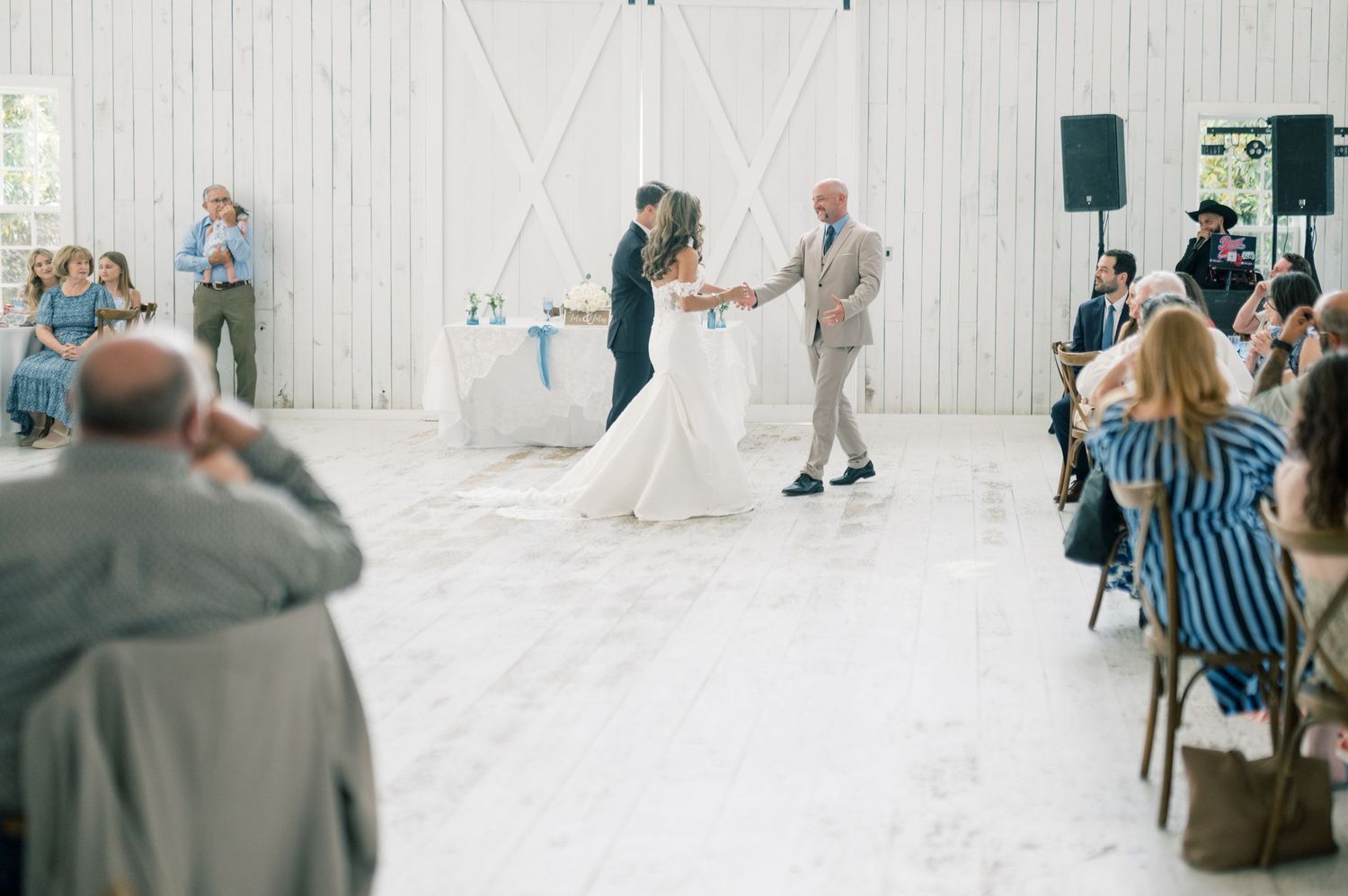 Bride and groom sharing their first dance in a white barn venue surrounded by seated wedding guests.