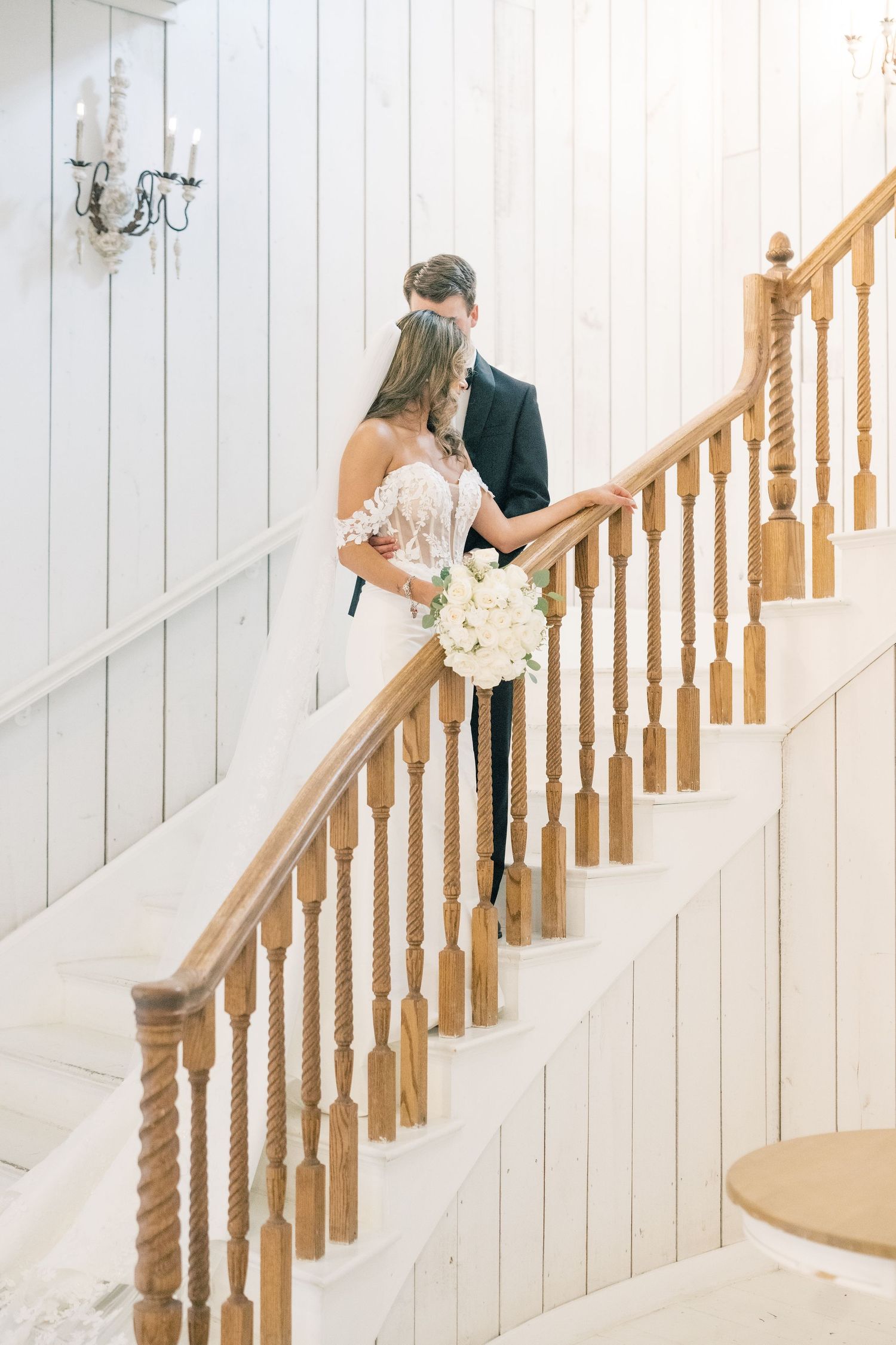 Bride and groom sharing a romantic moment on a white wooden staircase with oak railings.
