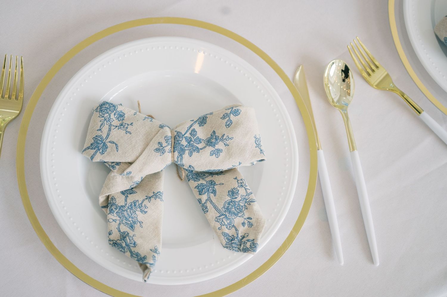 Elegant white plate with gold rim charger, gold cutlery, and blue floral patterned bow napkin.