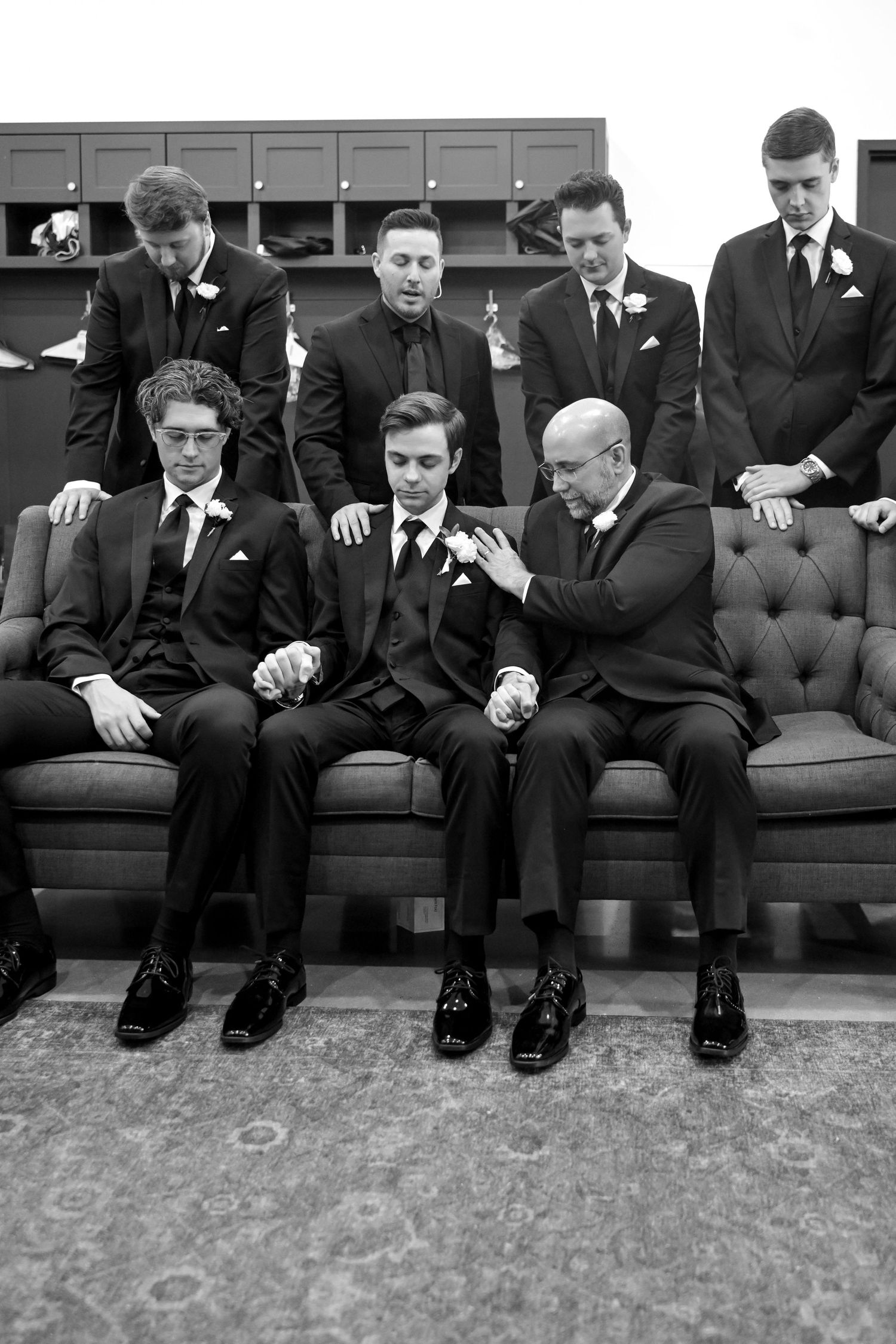 Groomsmen in black suits gather on a tufted sofa for a prayer before the wedding ceremony.