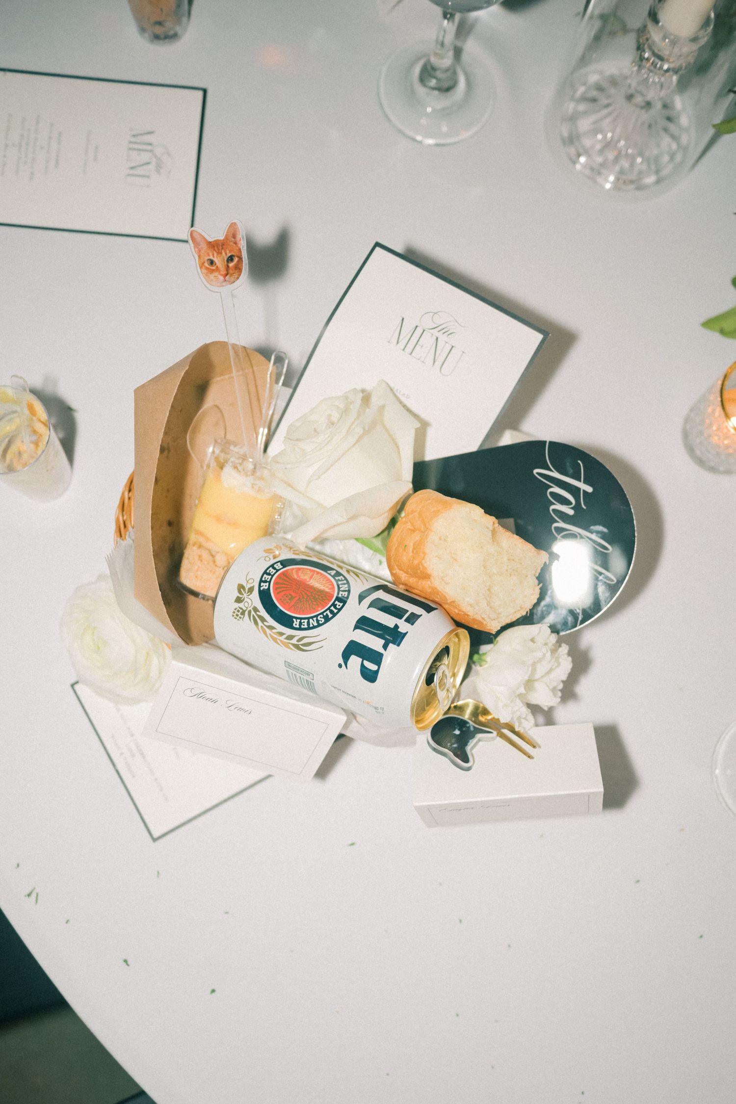 Wedding reception table detail with Miller Lite can, bread roll, menu card, and white roses.