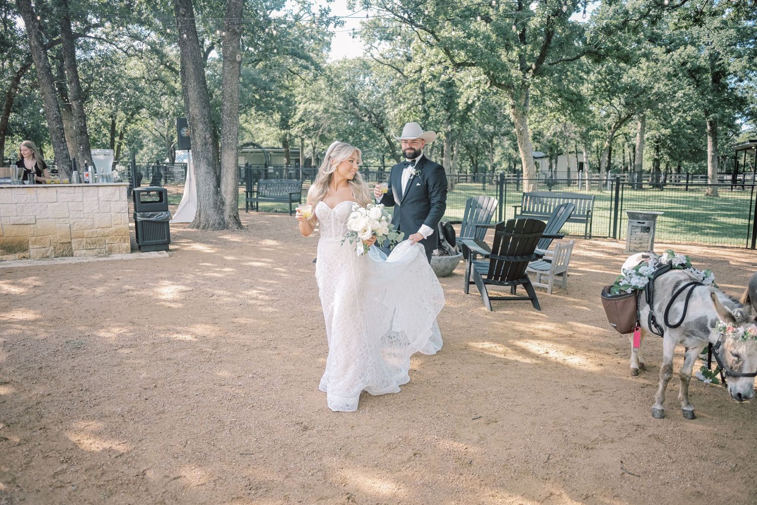 Bride and groom walk through tree-lined outdoor venue with a decorated donkey nearby.