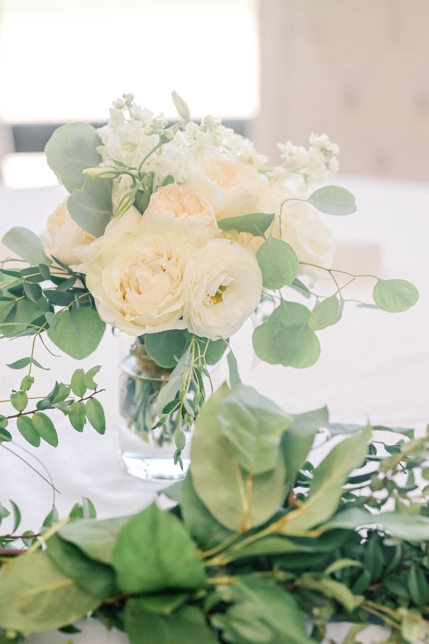 Elegant white peony and rose centerpiece with eucalyptus greenery in glass vase on white tablecloth.