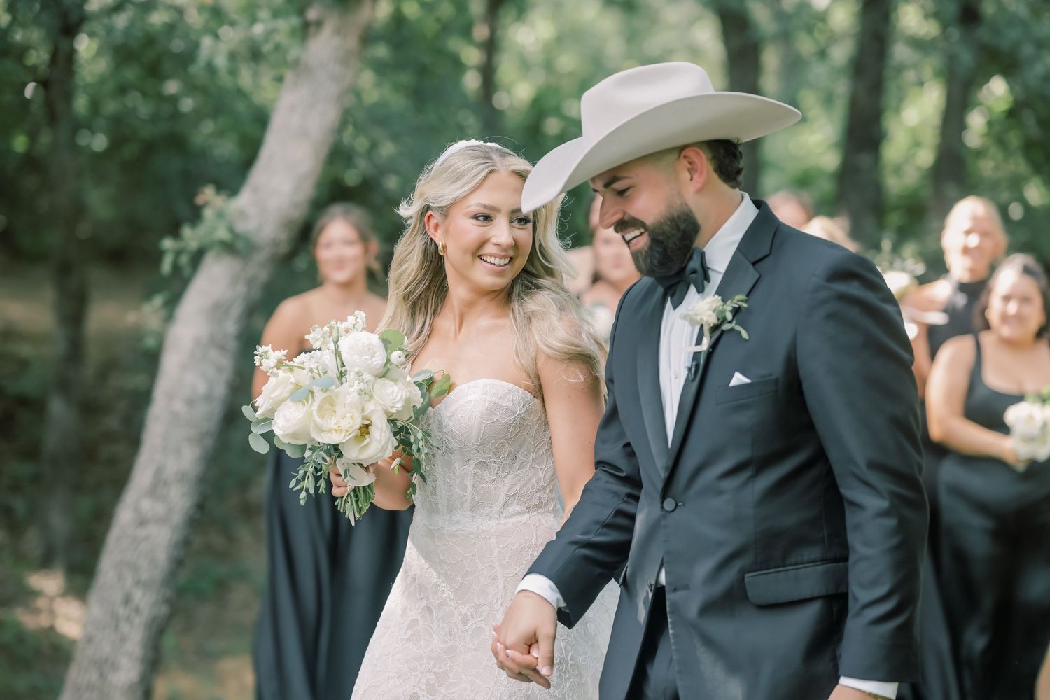 Smiling bride and groom in cowboy hat walking hand in hand after outdoor wedding ceremony.