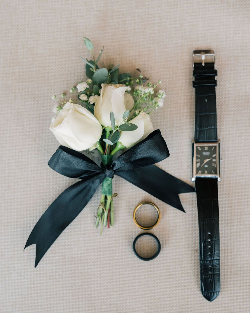 Wedding flat lay with white rose boutonniere, black ribbon, watch, and gold and black rings.