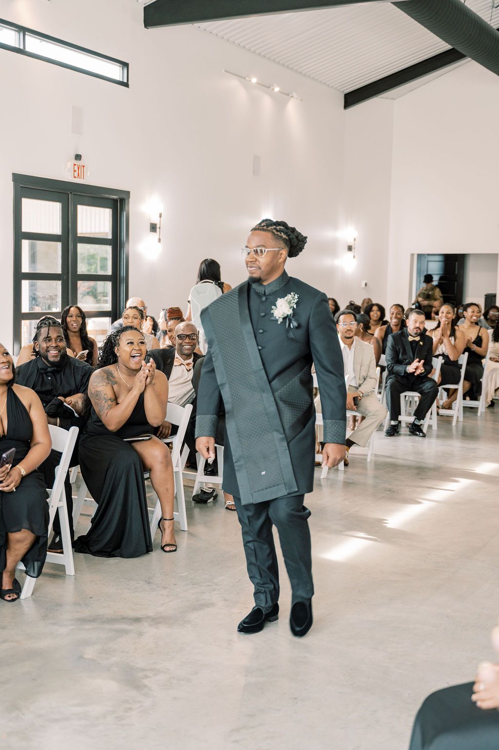 Groom walks down the aisle in a dark teal suit with boutonniere as wedding guests smile and applaud.