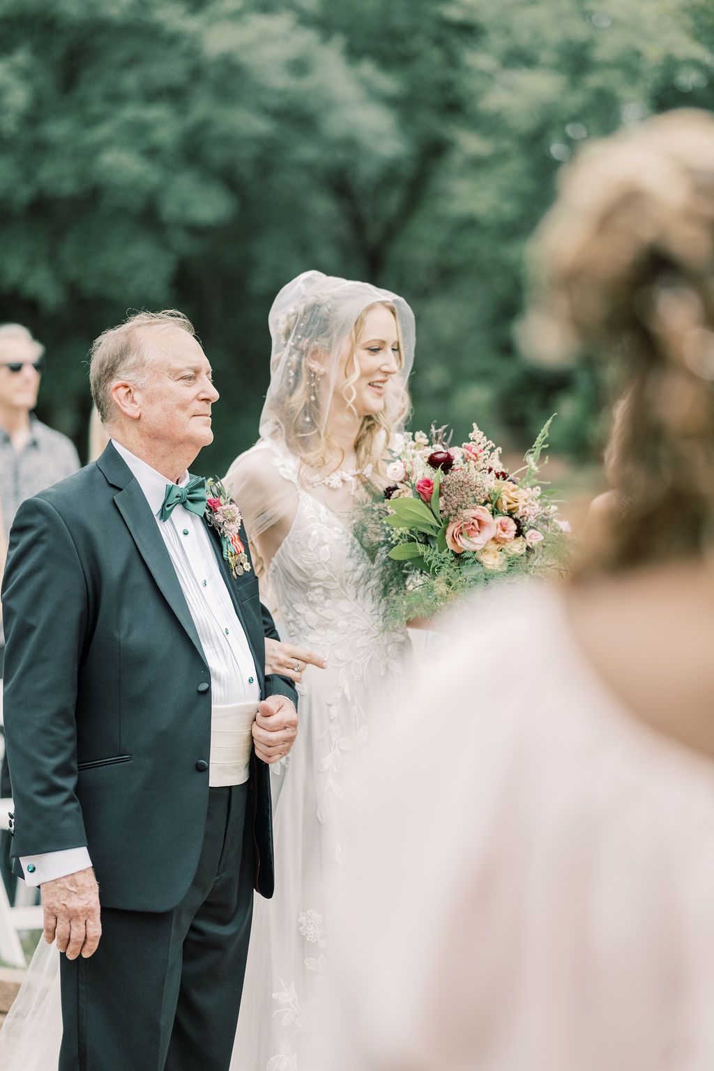 Bride in lace gown and veil holds colorful bouquet while walking down the aisle with an older gentleman in a dark suit.
