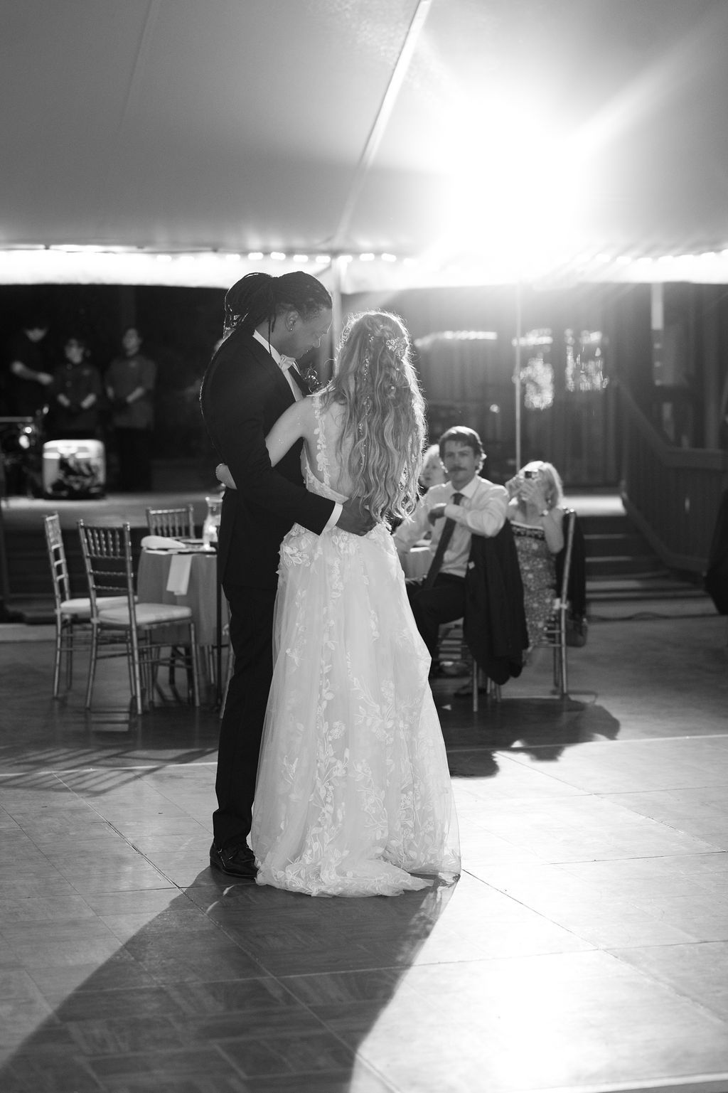 Black and white photo of bride and groom sharing first dance at wedding reception with guests seated in background.