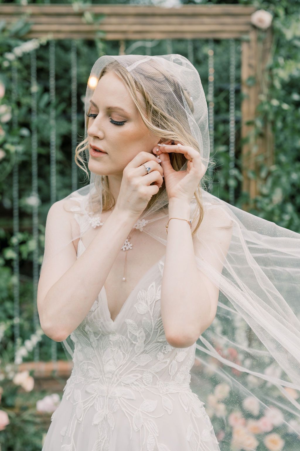 Bride in lace wedding gown adjusts earring under flowing veil, surrounded by greenery and florals.