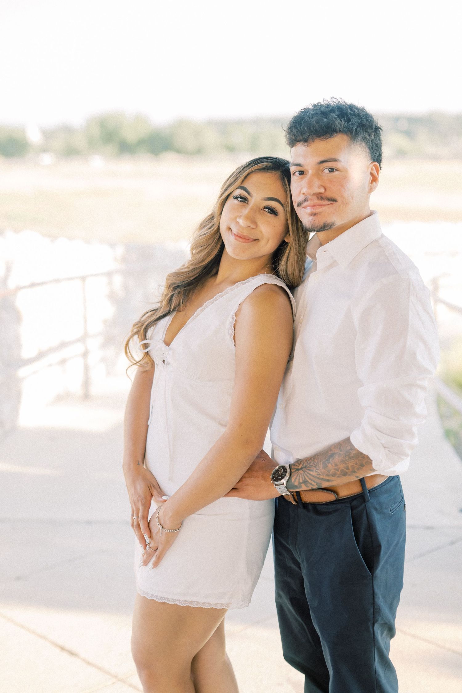A young couple poses outdoors, woman in white dress leaning back on man in white shirt and navy pants.