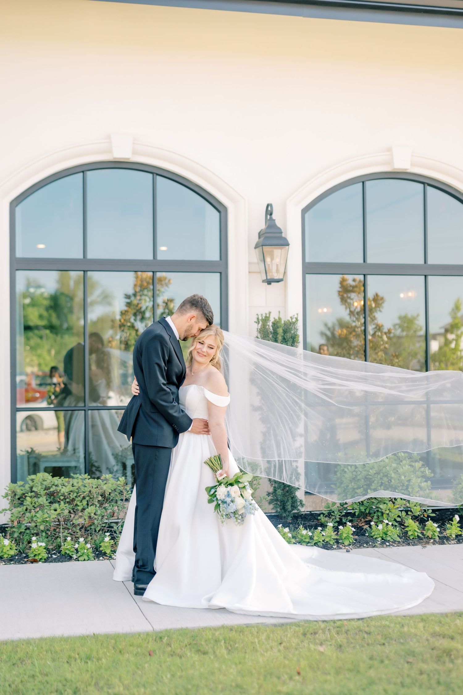 Bride in flowing white gown with veil blowing in wind embraces groom in navy suit outdoors.