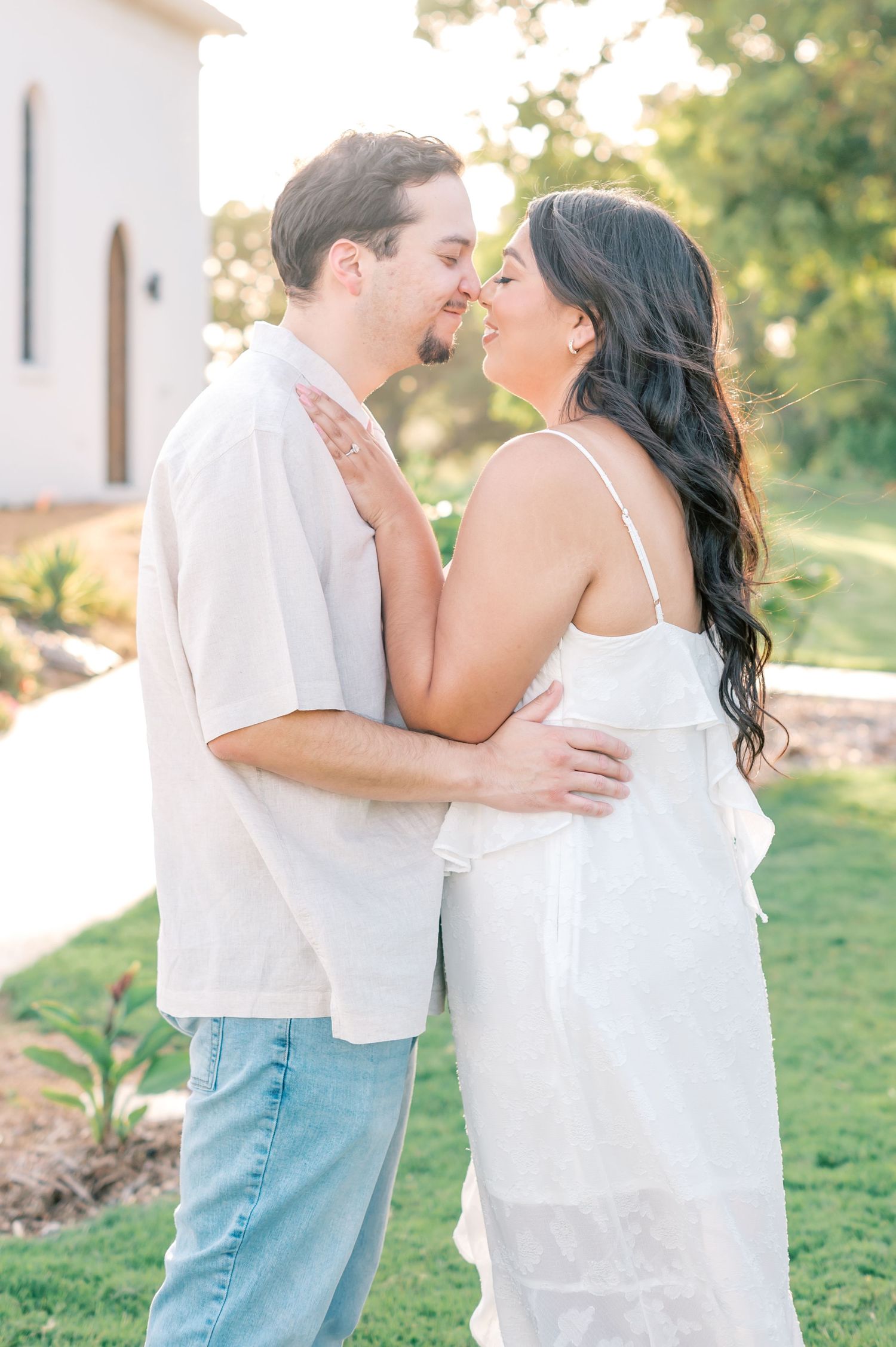 A romantic couple in white outfits share a tender moment outdoors with soft sunlight and greenery in the background.