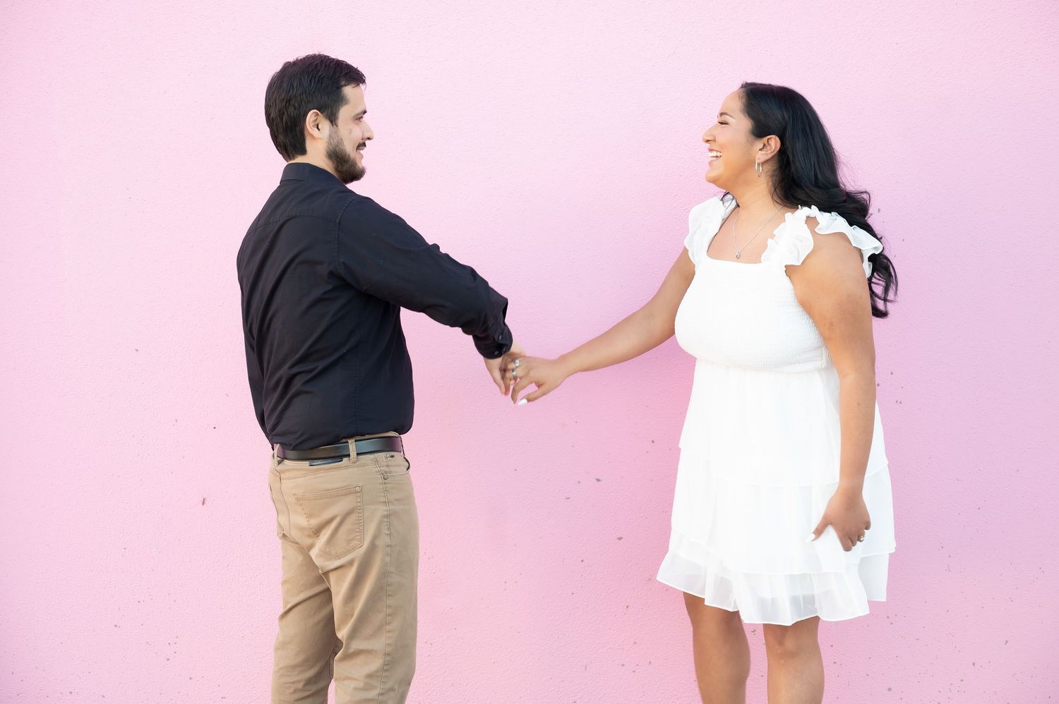 A couple holding hands and smiling against a pink wall during a photo shoot.