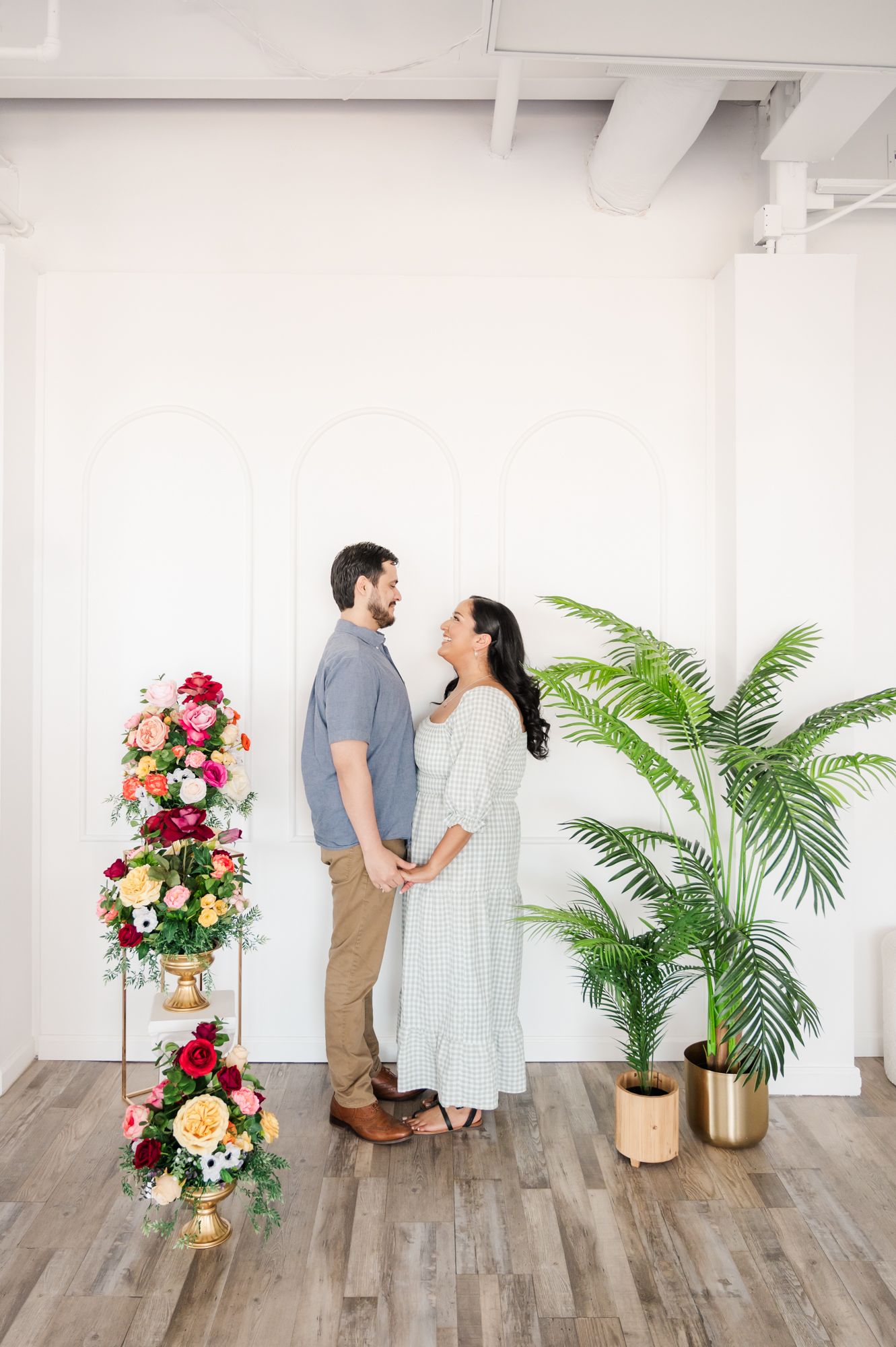 A romantic couple poses together in a bright white studio space with tropical plants and floral arrangements.
