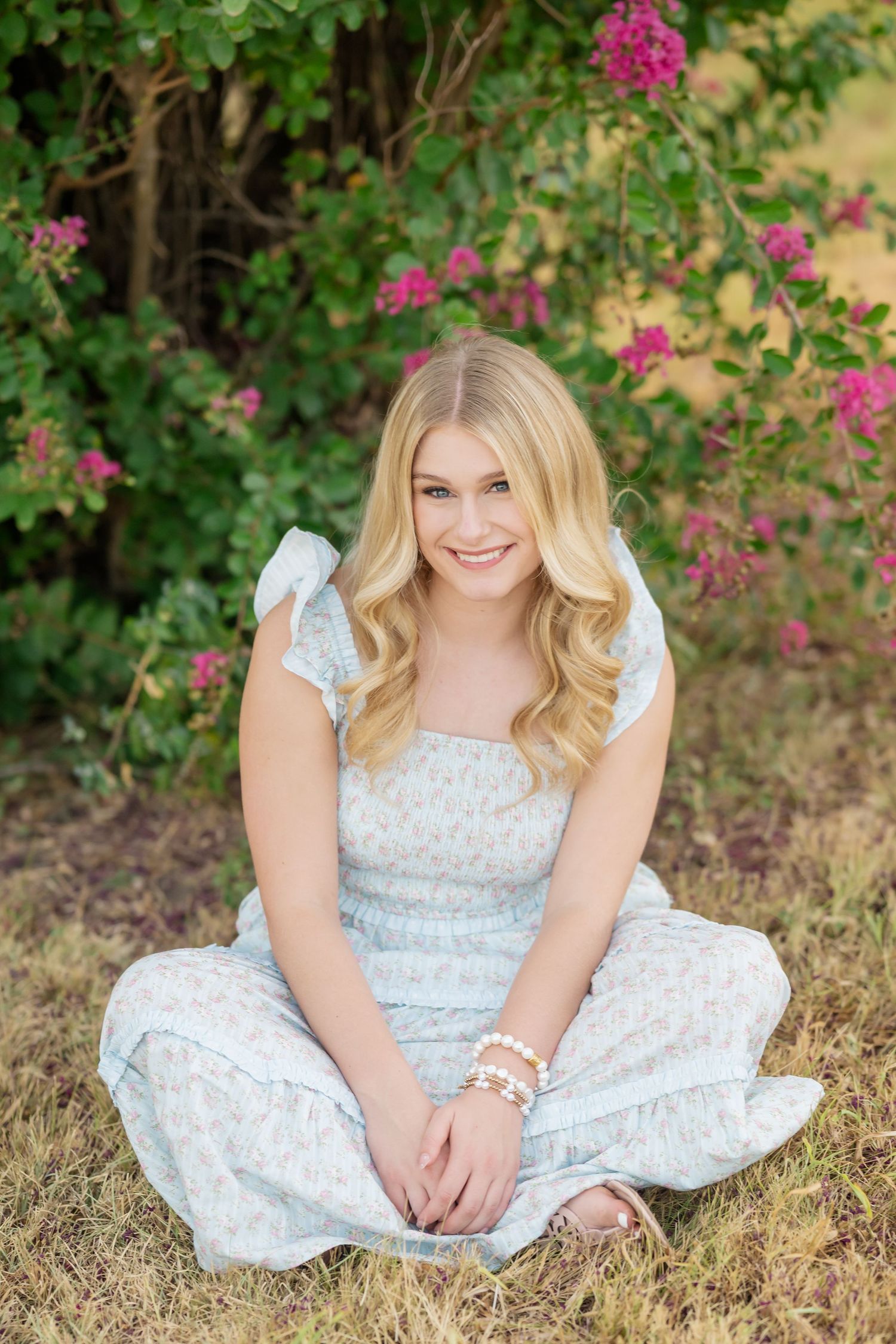 Smiling blonde teen girl in light blue floral dress sitting cross-legged on grass near pink flowering bushes.