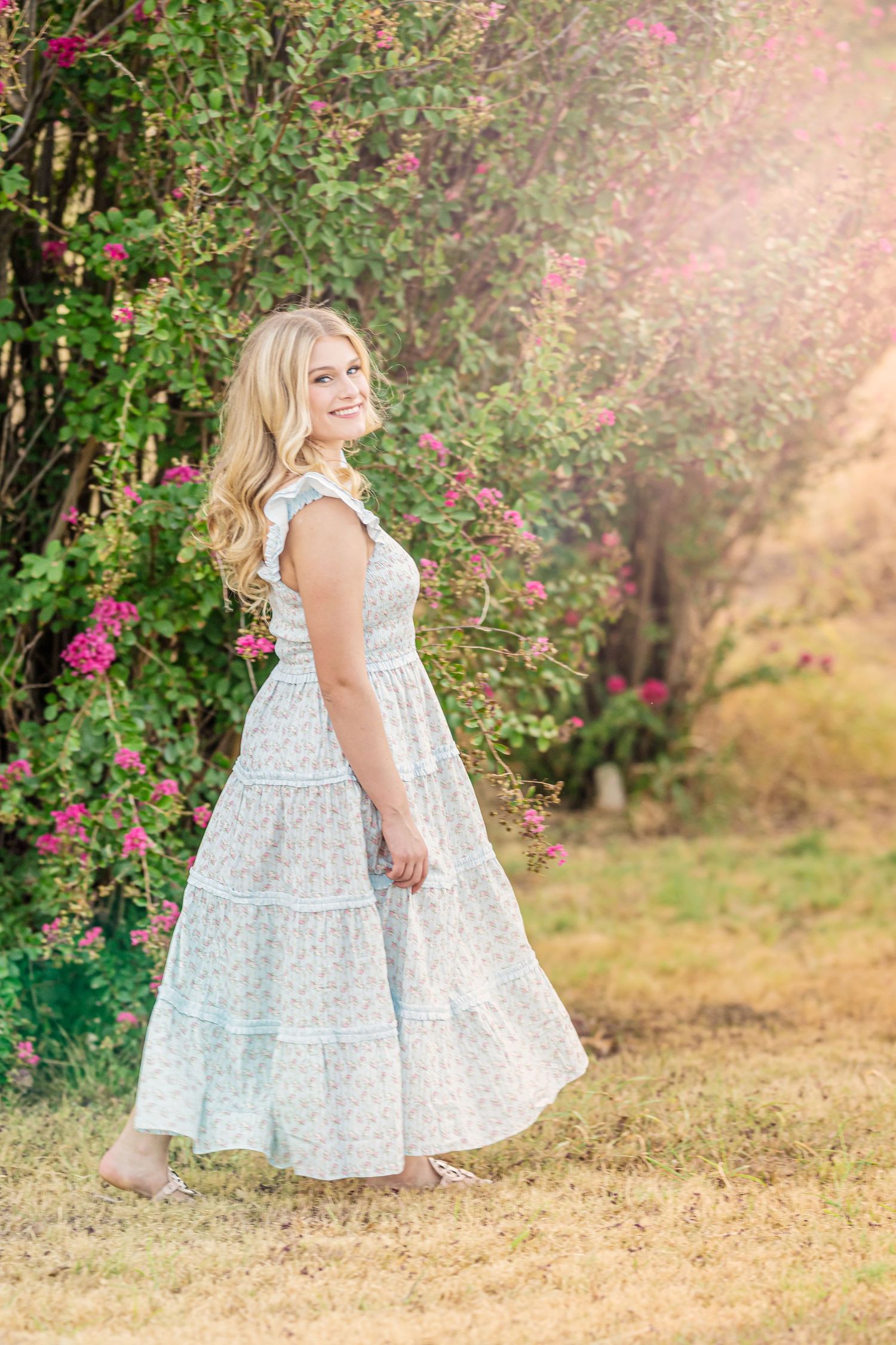 Smiling blonde woman in a floral maxi dress standing in a garden with pink blooming flowers on a sunny day.