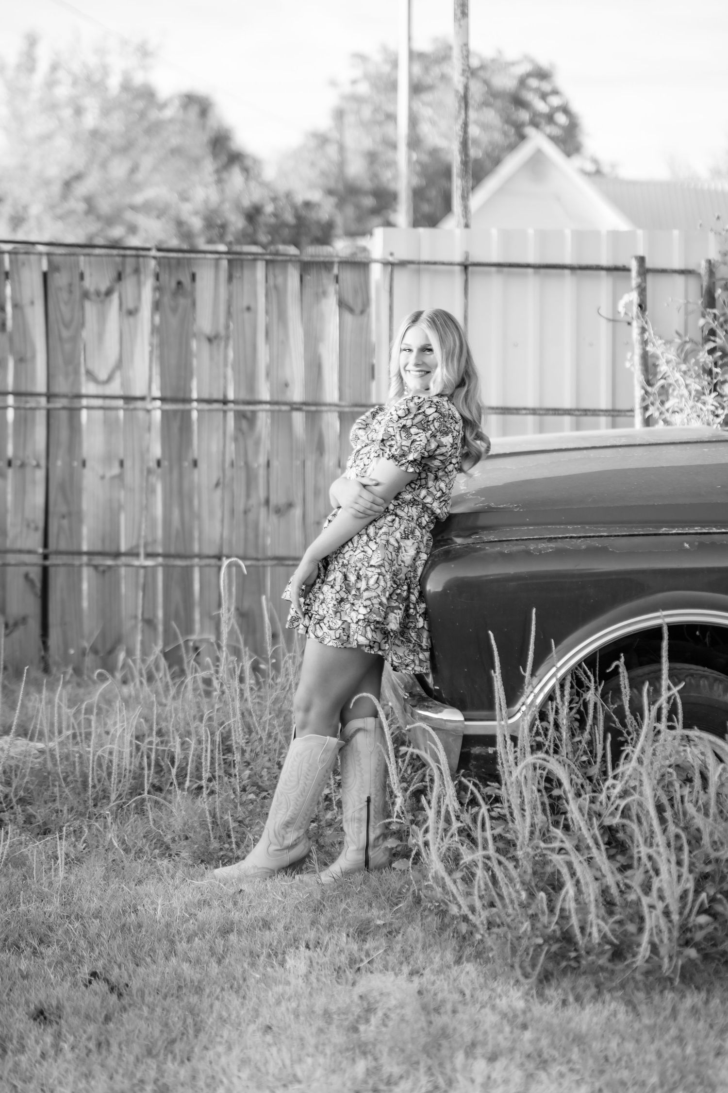 Black and white photo of a blonde woman in a floral dress and cowboy boots leaning against a vintage truck by a wooden fence.