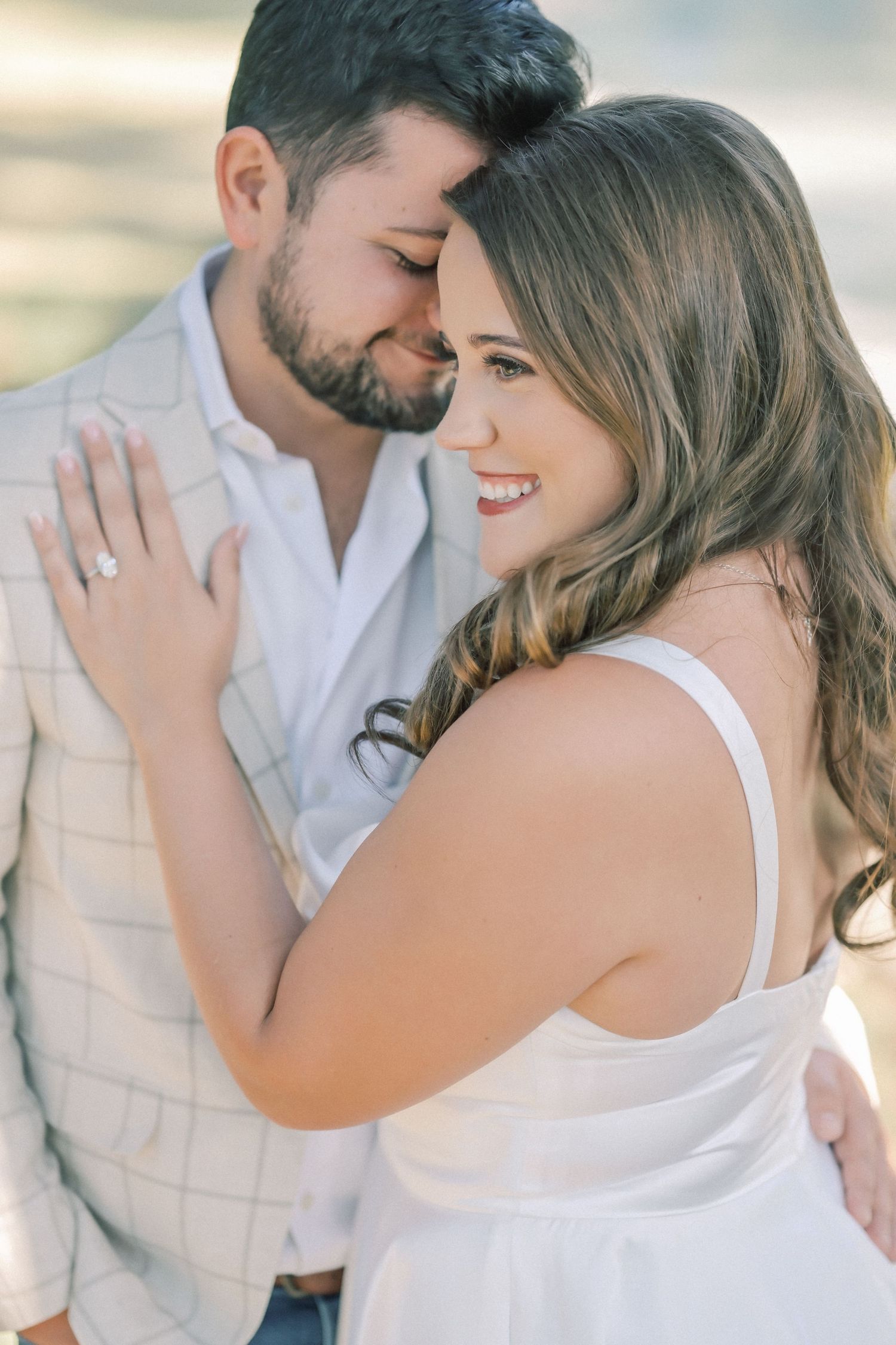Close-up engagement photo of a smiling woman showing her diamond ring as her bearded fiancé rests his forehead against hers.