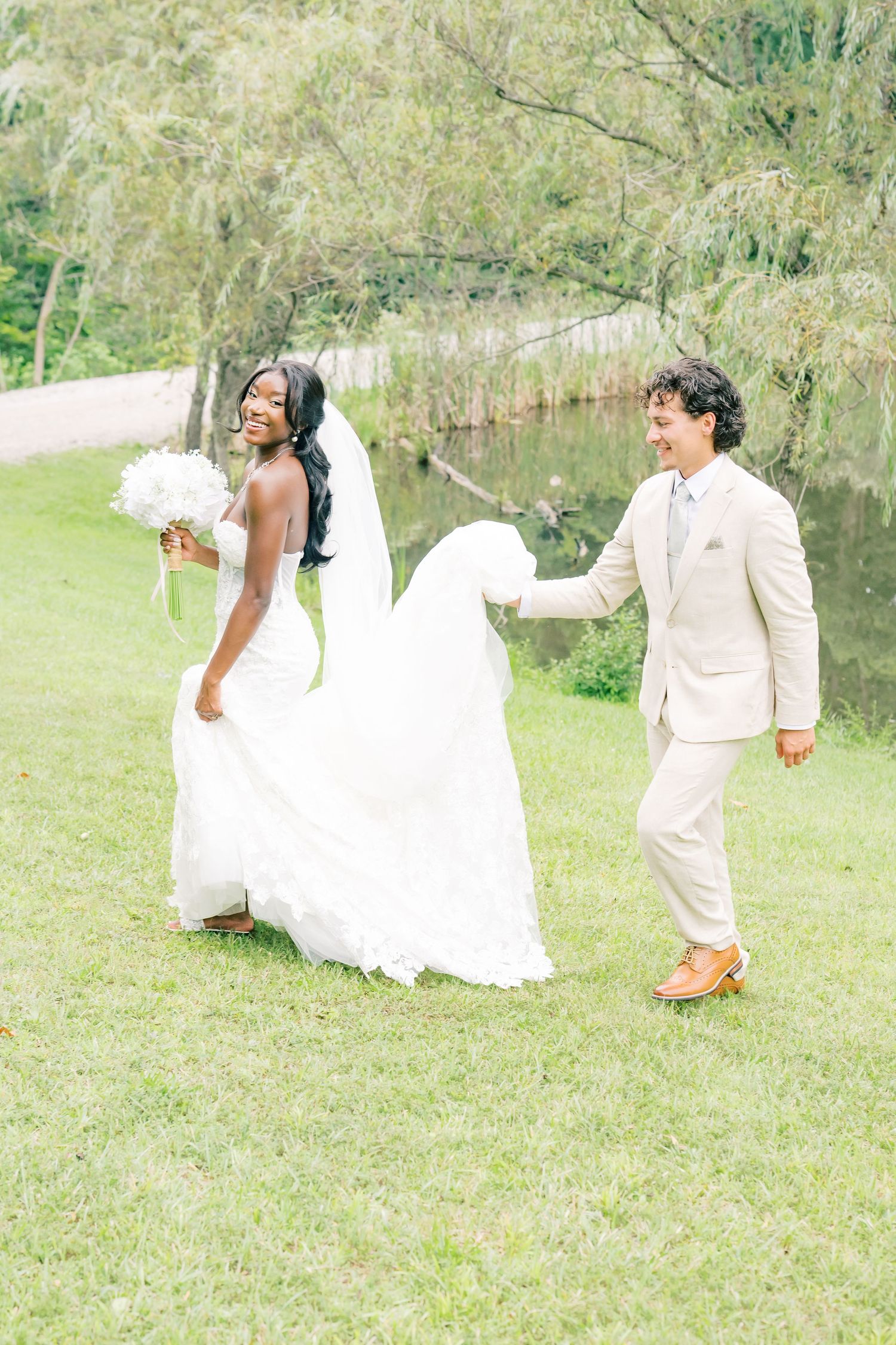 Joyful bride in lace gown holding bouquet while groom carries her train across lush green lawn by pond.