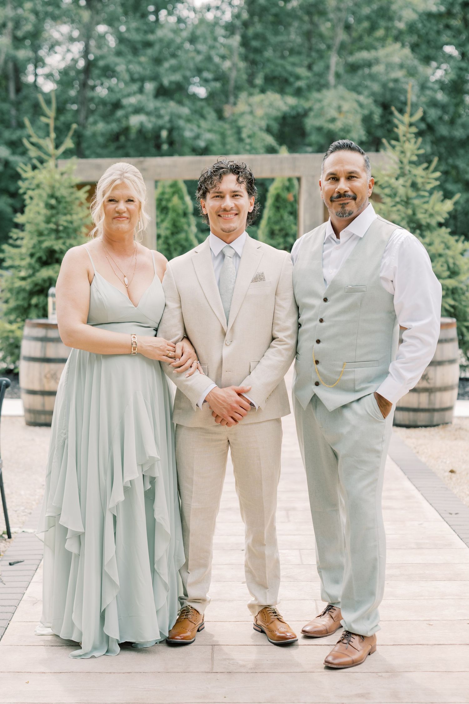 Groom in tan suit poses with bridesmaid in sage dress and groomsman at outdoor wedding venue.