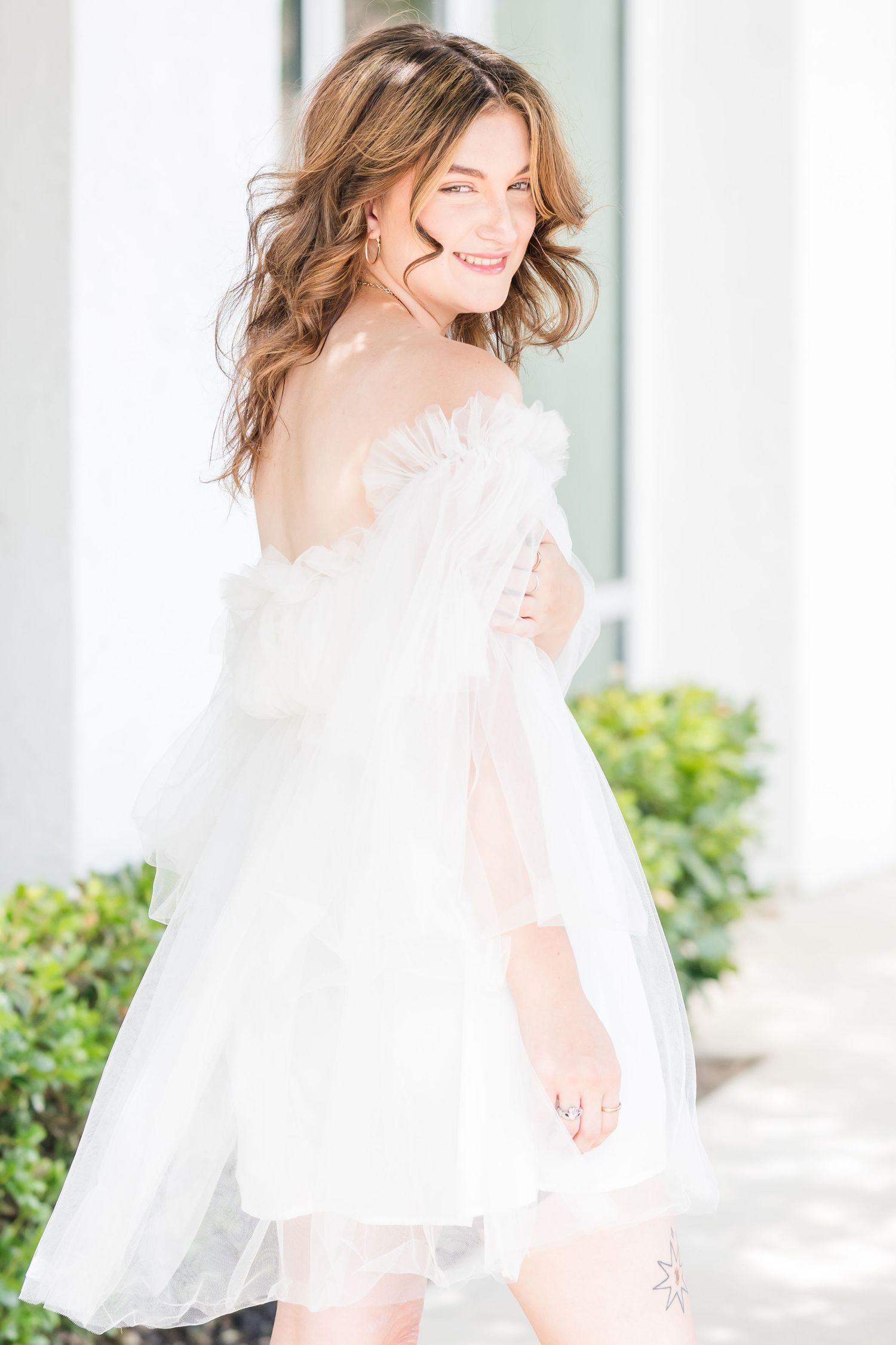 A smiling woman in a white off-shoulder tulle mini dress poses outdoors near white columns and green shrubs.