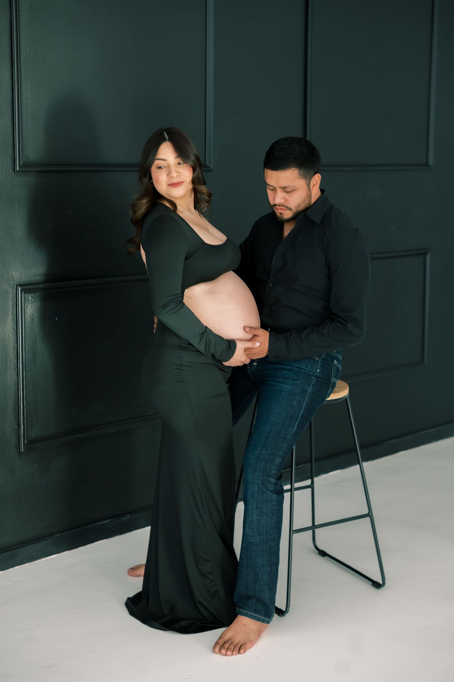 Couple in dark green outfits posing for maternity photo, man seated on stool touching pregnant woman's belly.