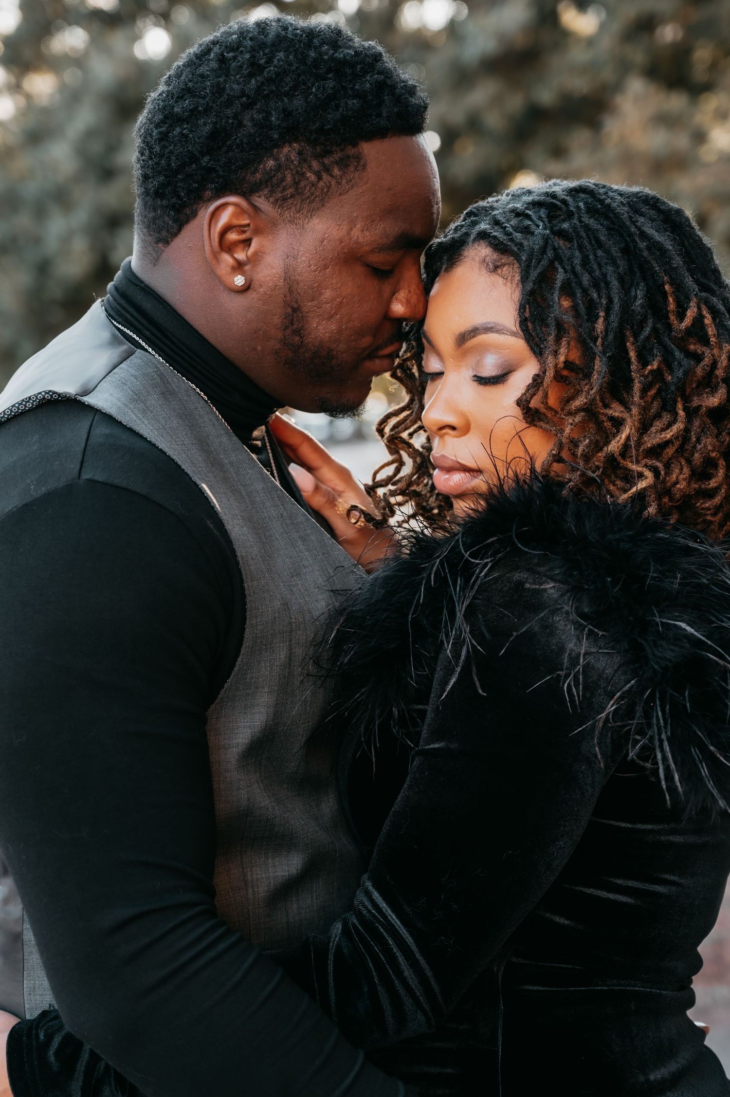 A romantic couple in elegant black outfits sharing an intimate moment, their foreheads touching tenderly outdoors.