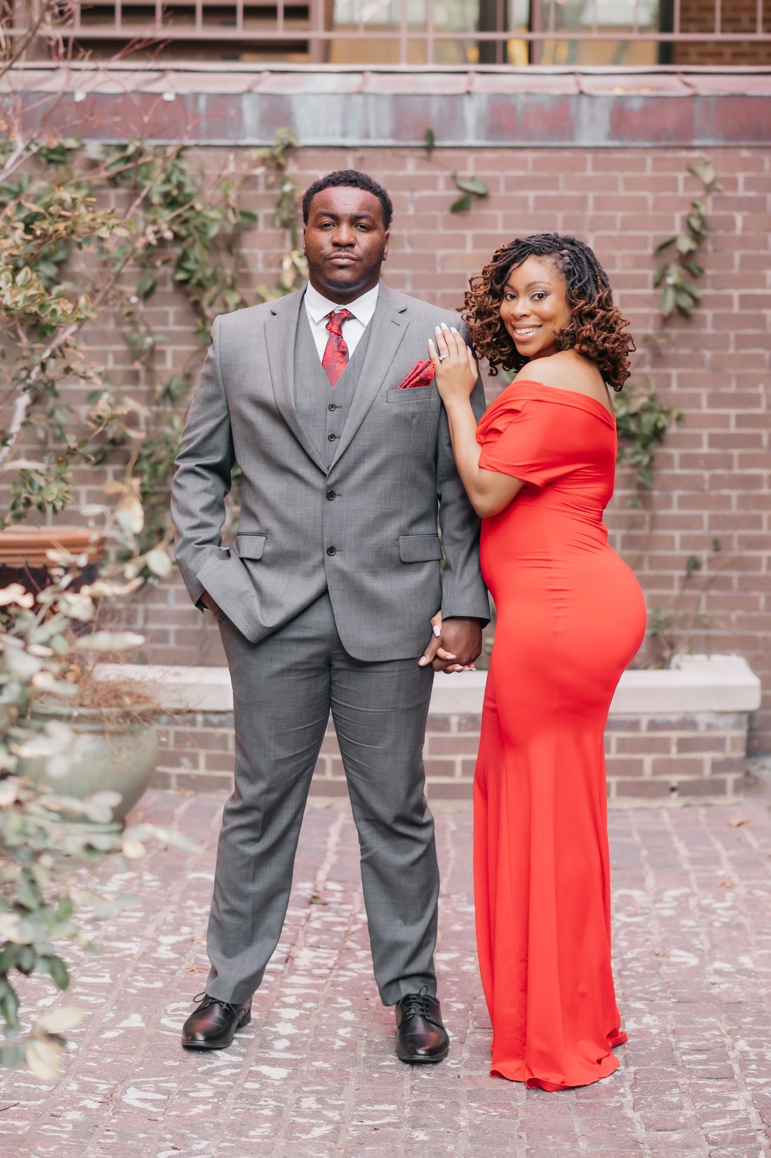 Stylish couple holding hands outdoors, man in gray three-piece suit and woman in fitted red dress.