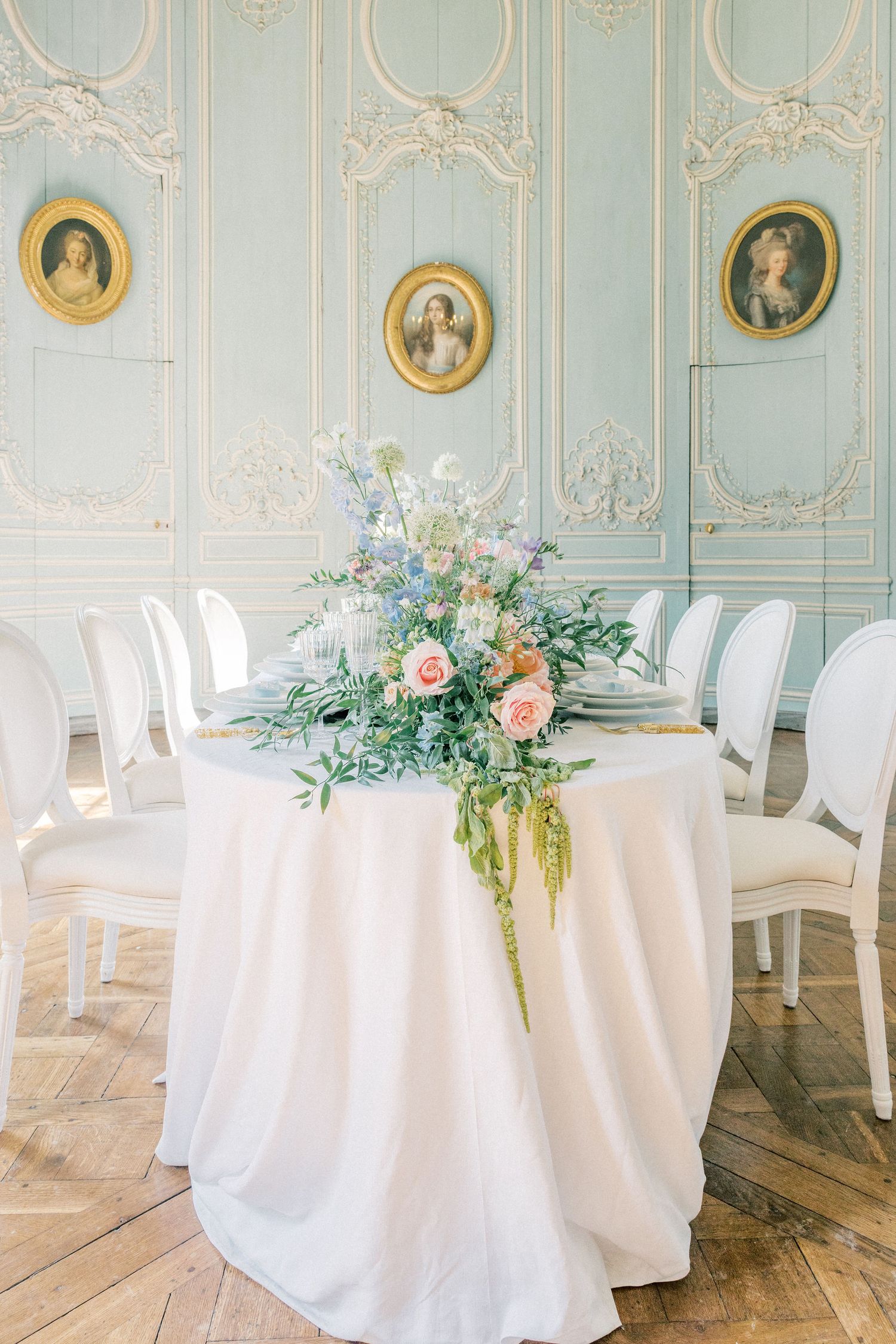 Elegant wedding reception table with pastel floral centerpiece in a French-style room with blue ornate wall panels.