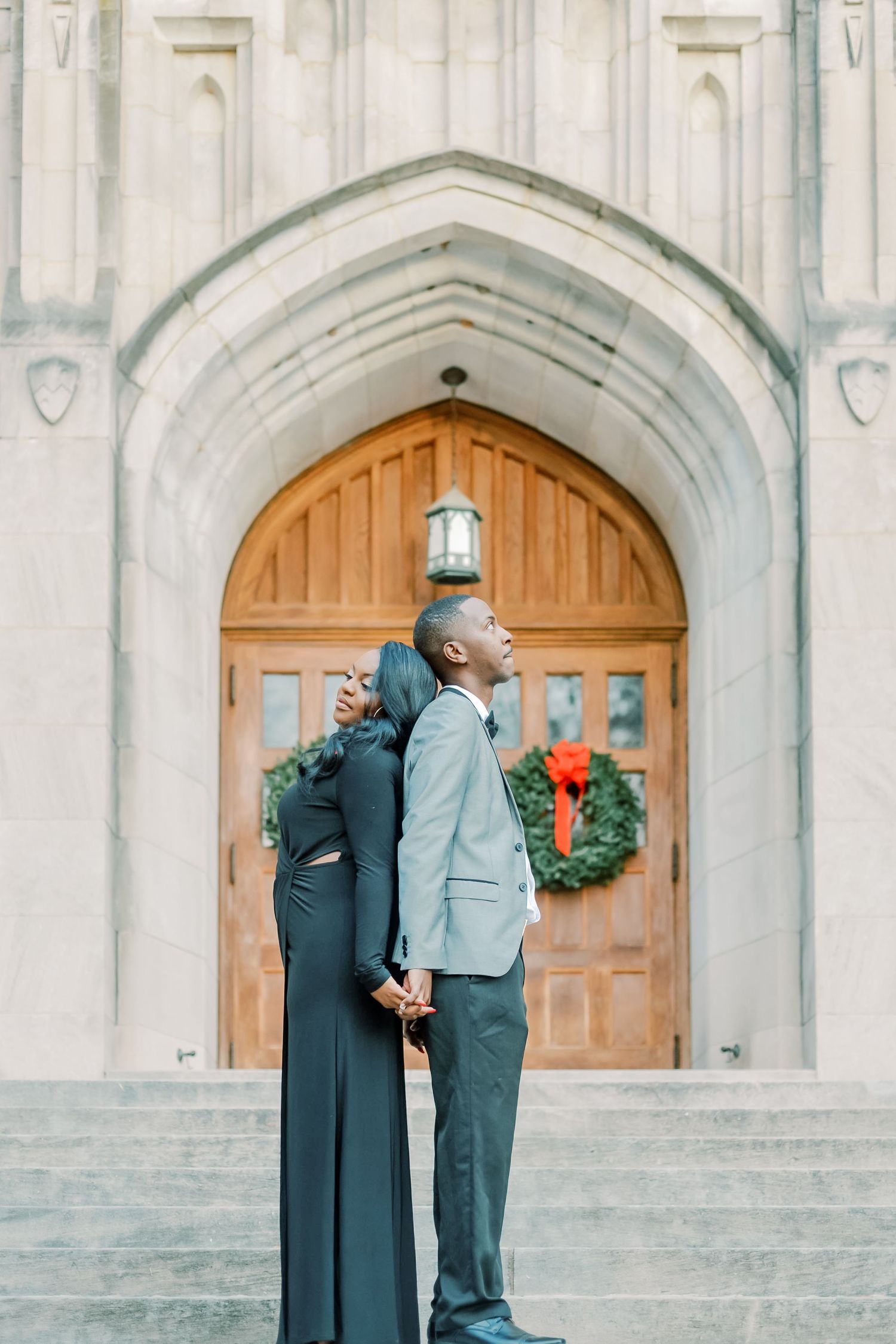Elegant couple in formal attire standing back-to-back on church steps with holiday wreath backdrop.