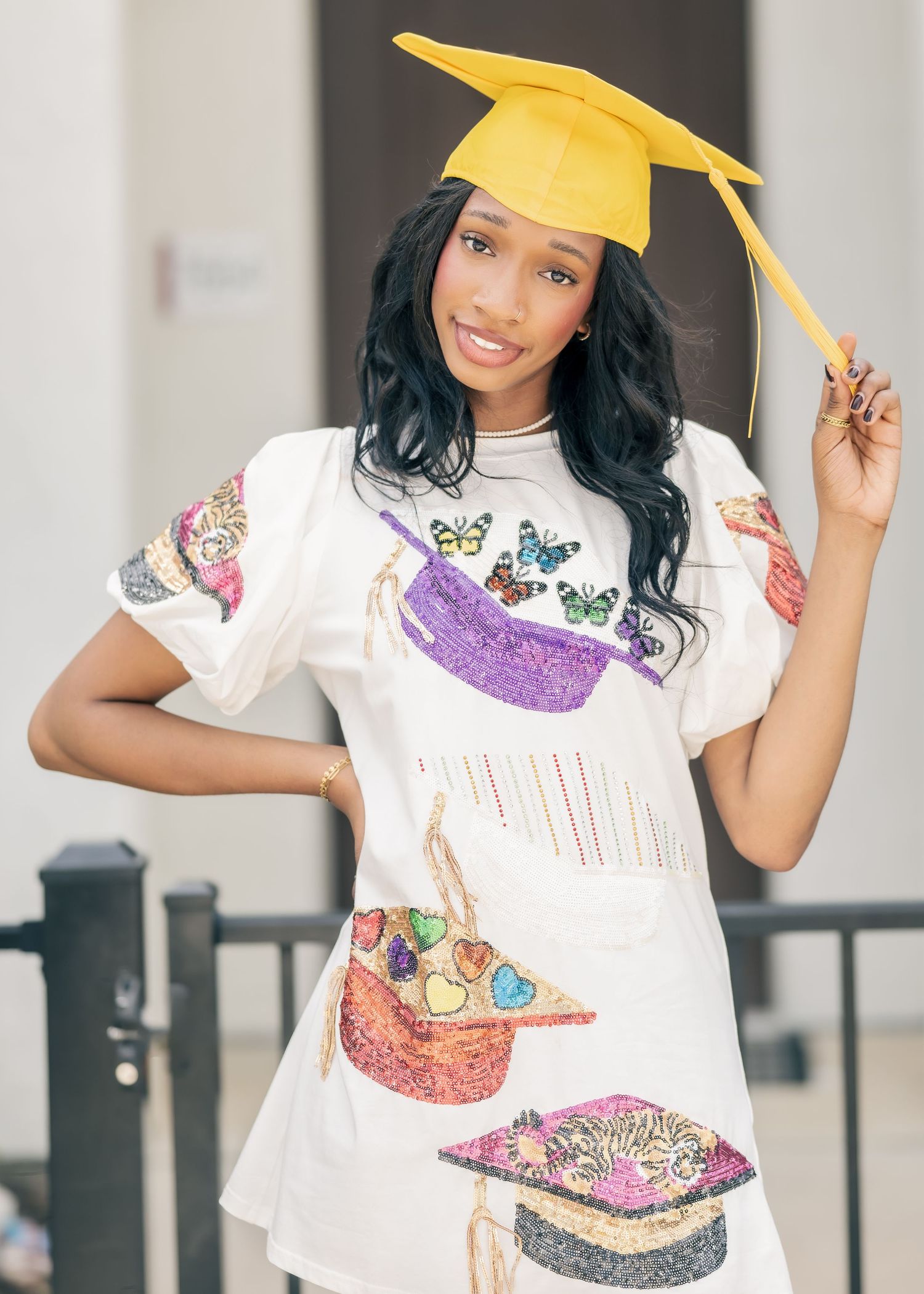 Young woman in a graduation cap and white dress with colorful sequined mortarboard prints poses for photos.