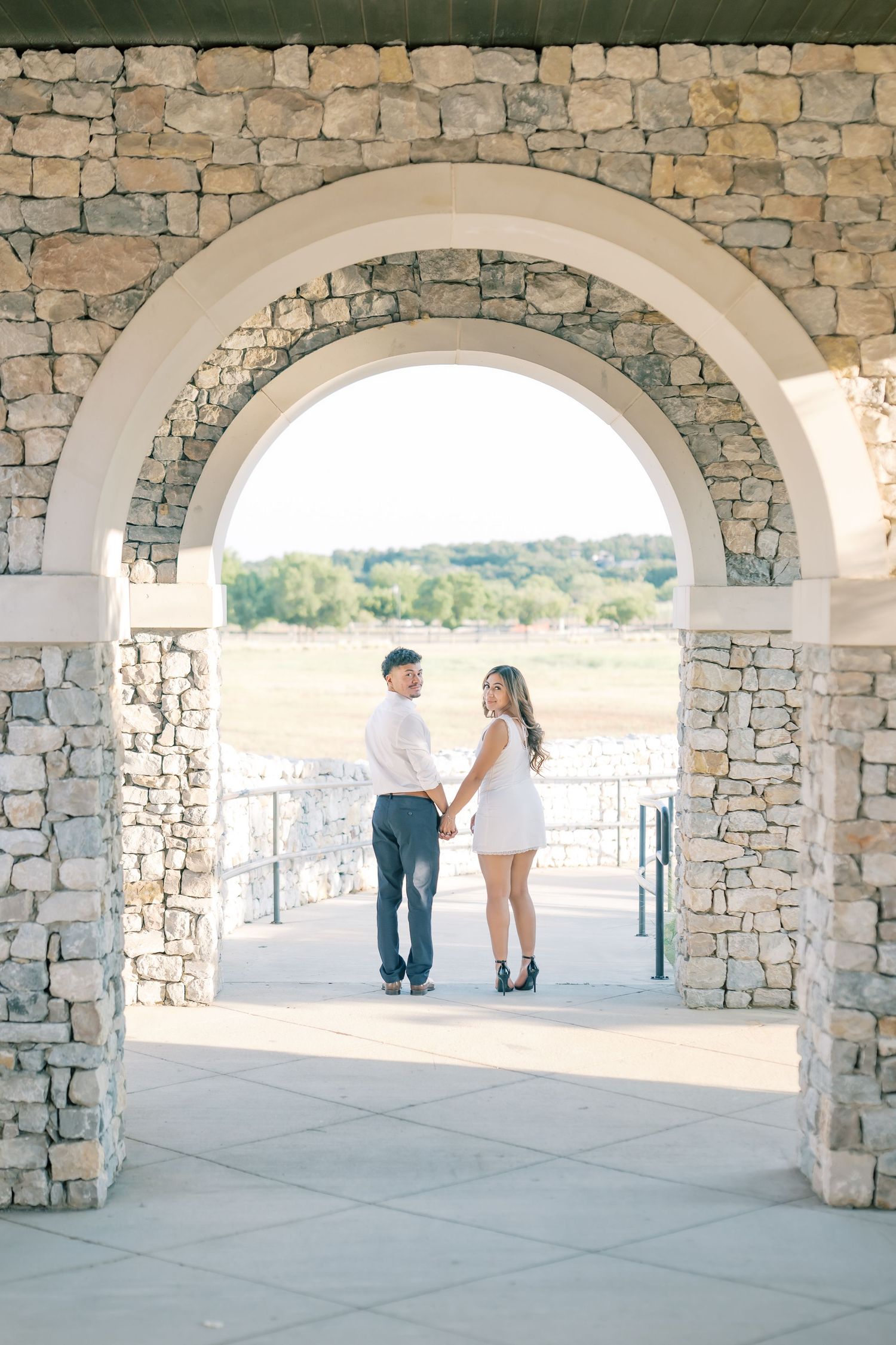 Couple holding hands framed by a stone archway during golden hour engagement photography session.