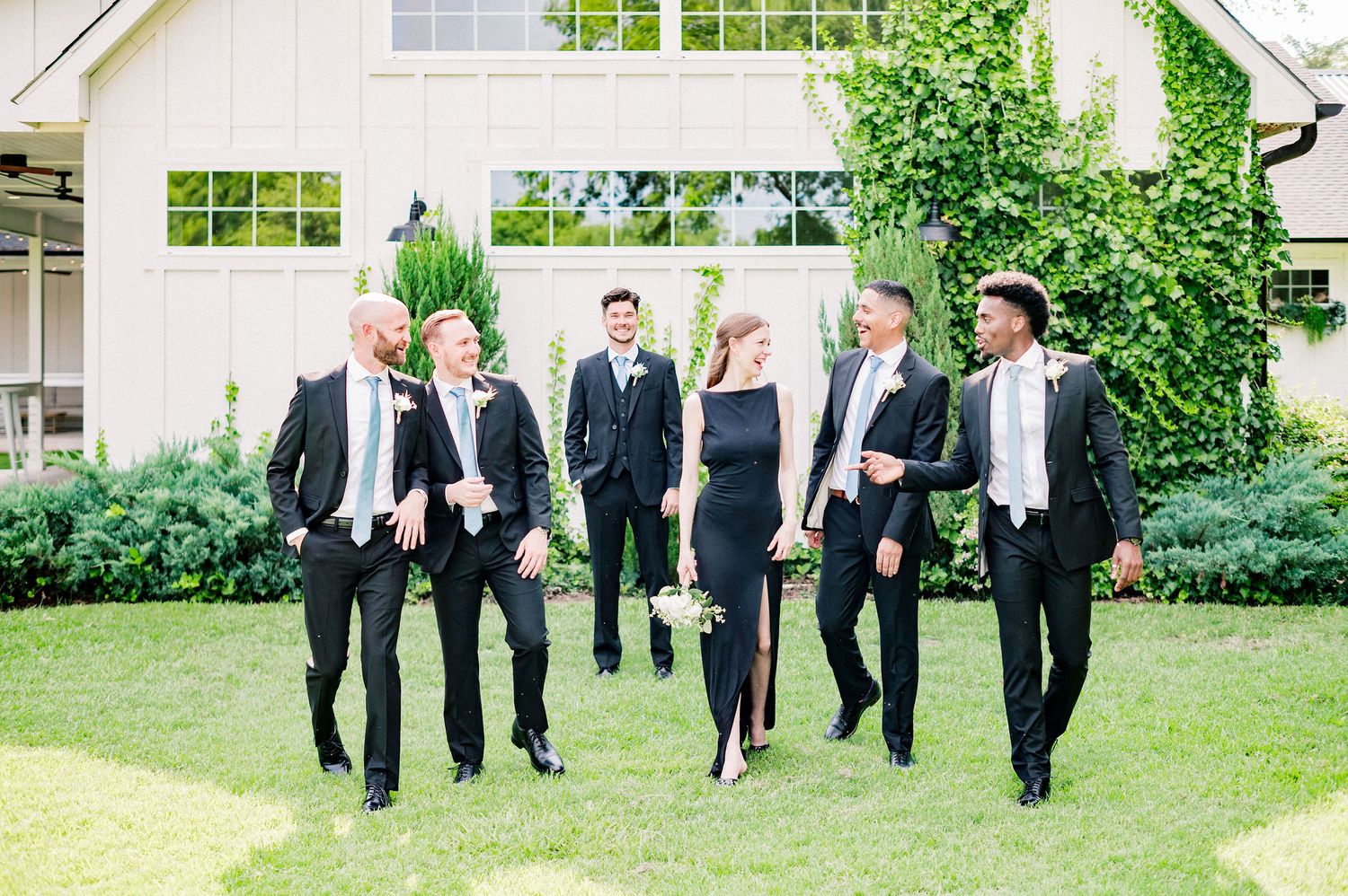 Wedding party in formal black attire walking and laughing together on a grassy lawn outside a white building.