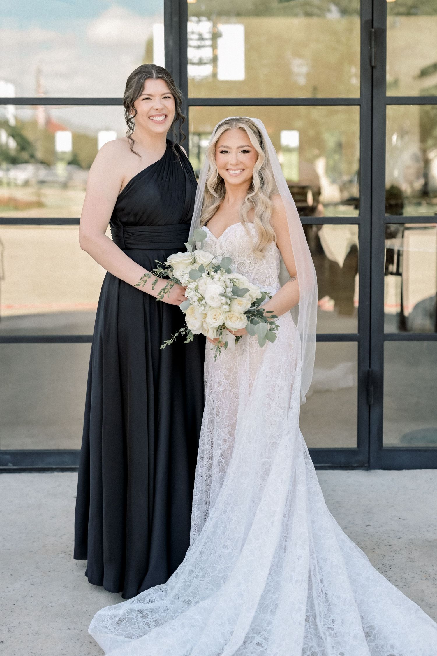 Bridesmaid in black gown poses with smiling bride in lace wedding dress and veil.