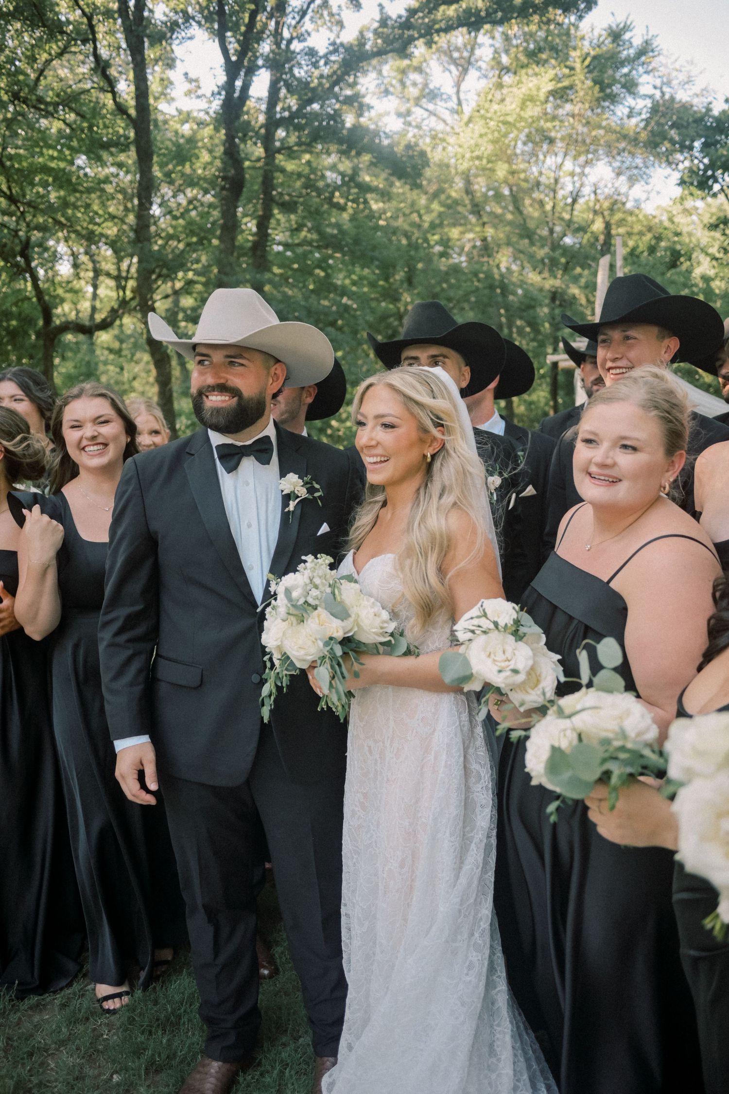 Bride and groom with wedding party, groomsmen in black cowboy hats, bridesmaids in black gowns outdoors.