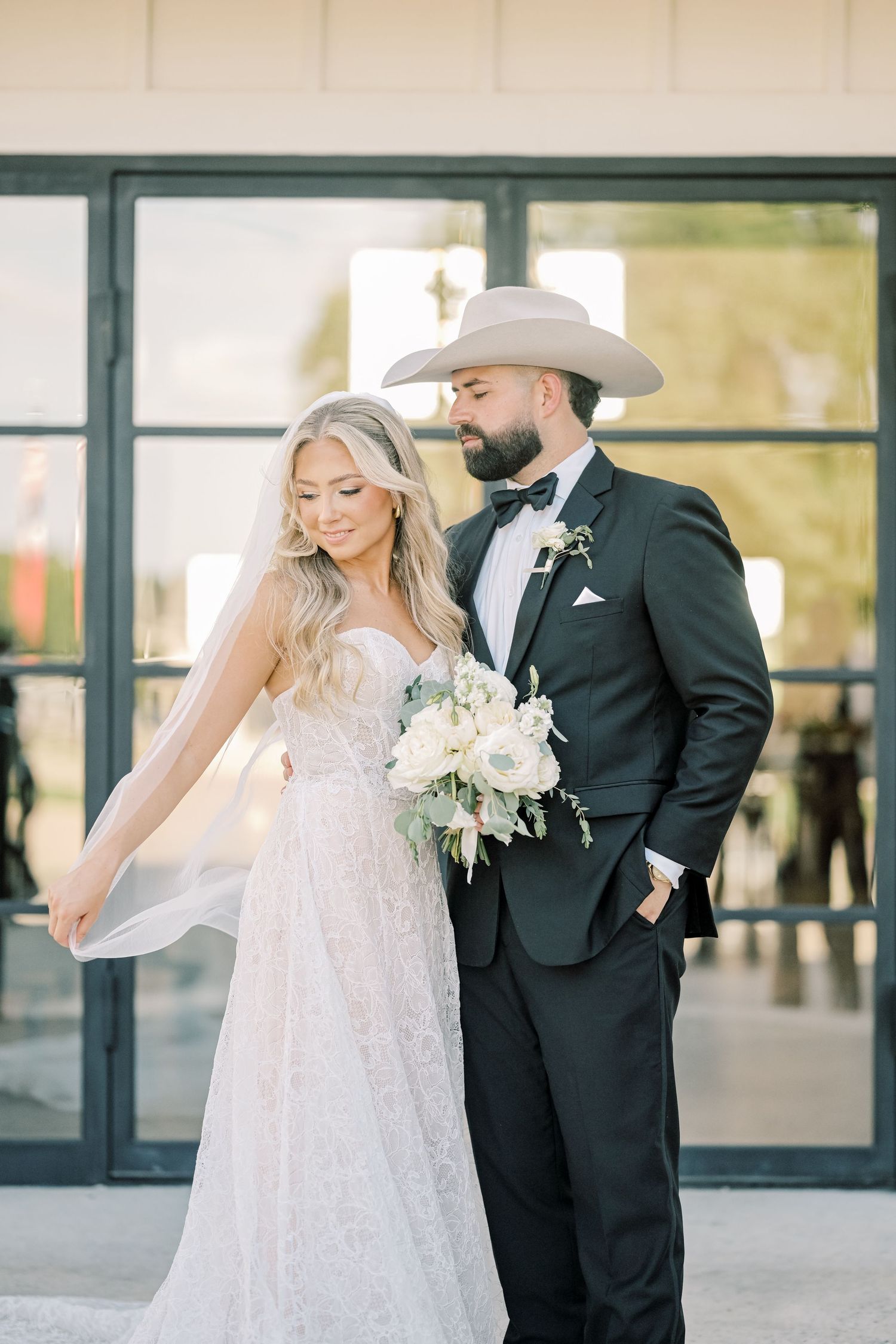 Bride in lace gown and groom in black tuxedo with cowboy hat pose with white bouquet.