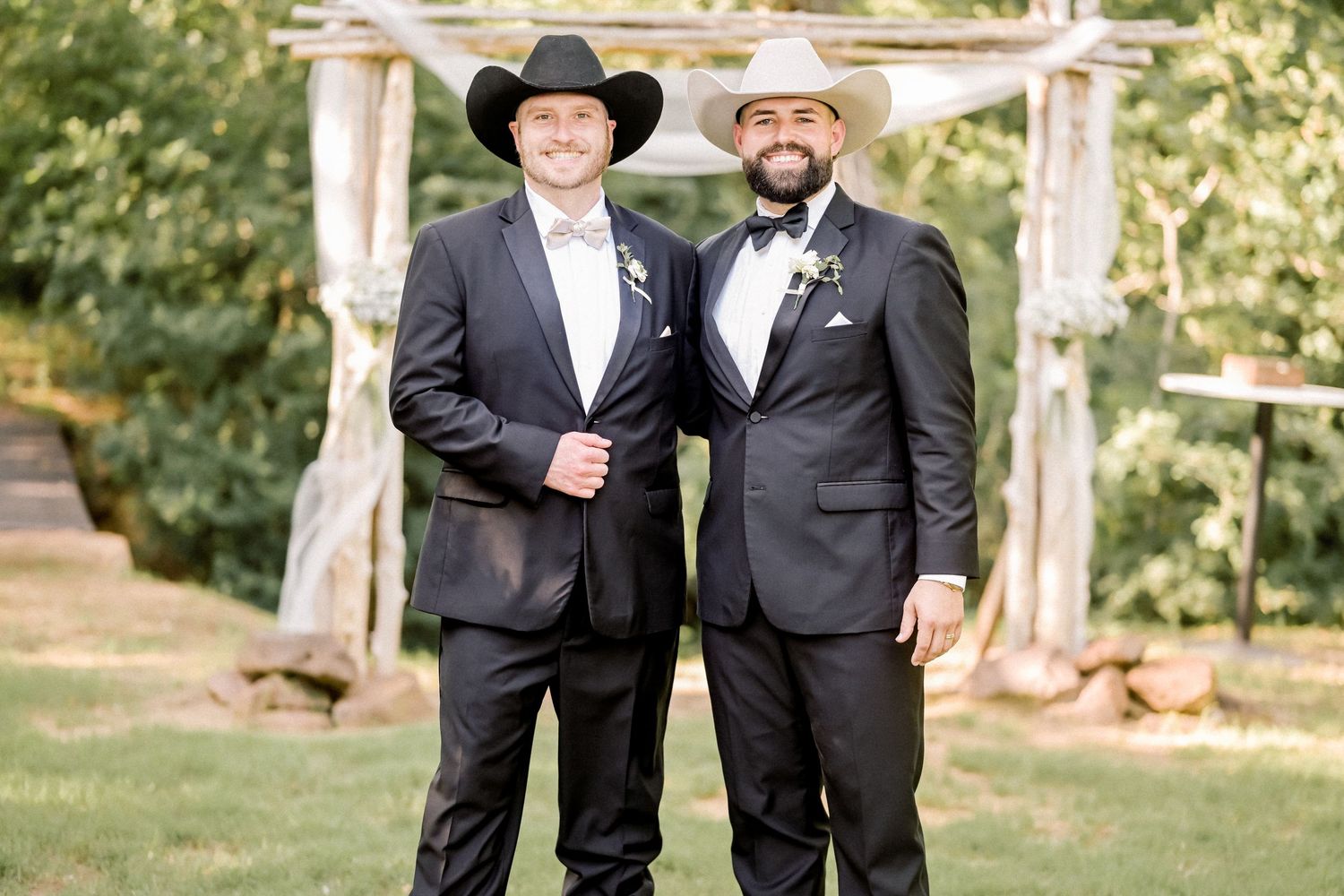 Groom and groomsman in black suits and cowboy hats pose before rustic wooden arch at wedding.