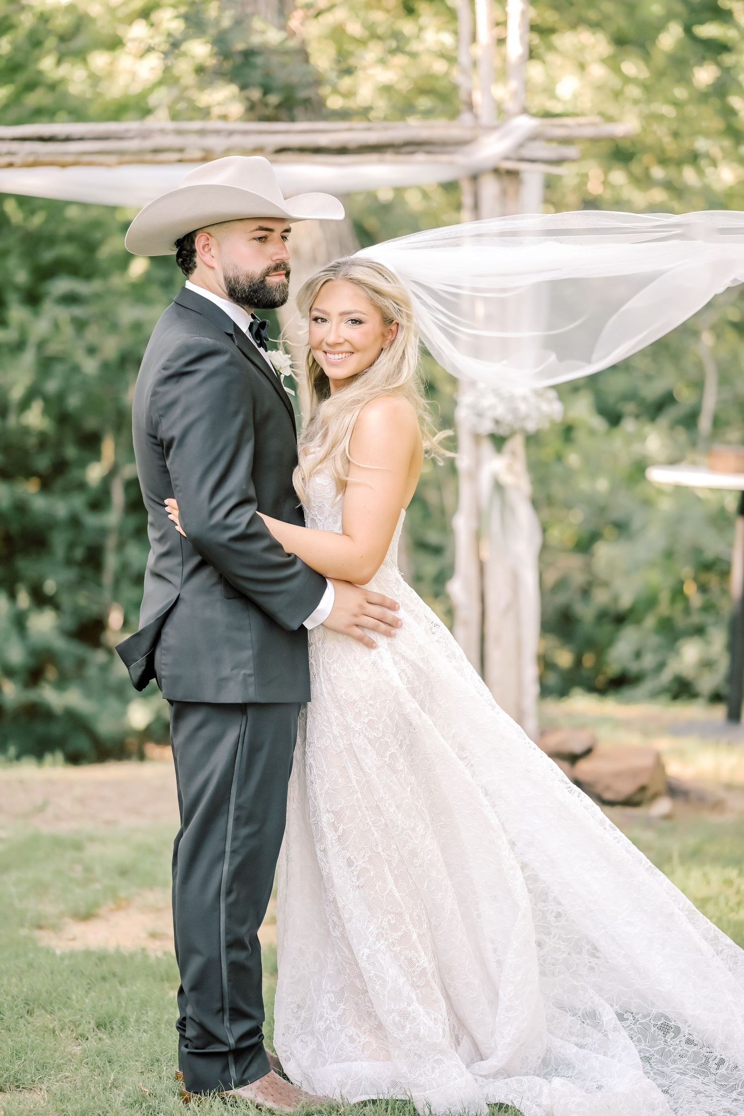 Bride in lace gown with flowing veil embraces groom in gray suit and cowboy hat at outdoor wedding.