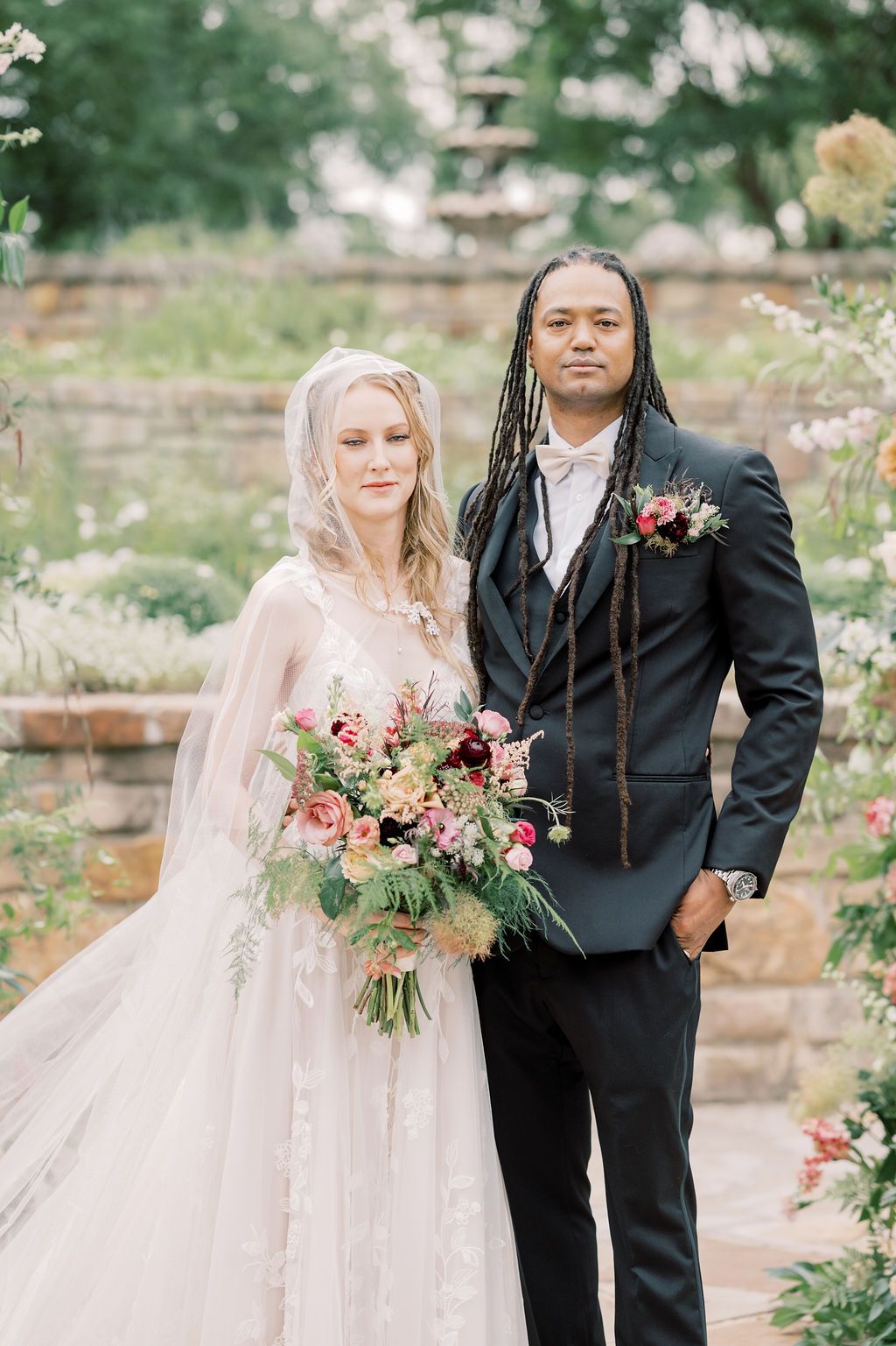 A bride in a lace gown with veil holds a floral bouquet beside a groom in a dark suit with dreadlocks outdoors.