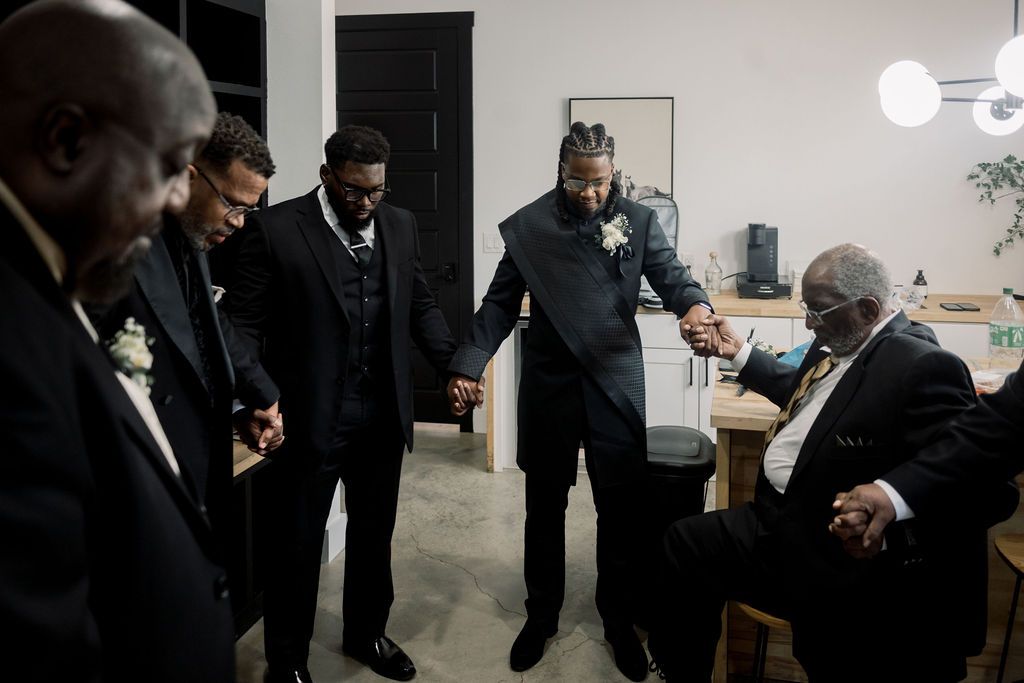Groomsmen in dark suits holding hands in prayer circle before wedding ceremony in dimly lit room.